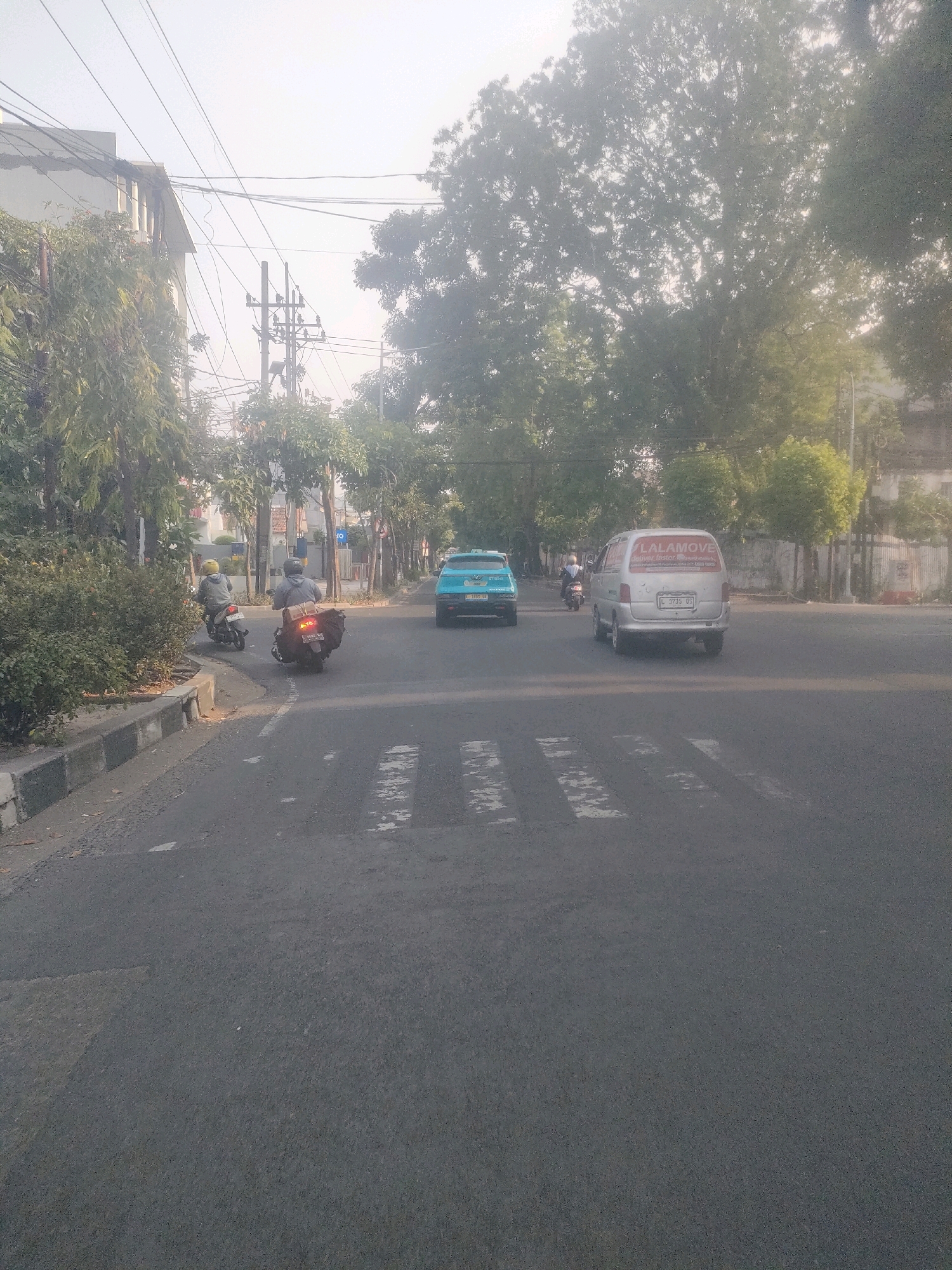 The image captures a street scene in Surabaya, Indonesia, likely in the early morning or late afternoon given the soft lighting and hazy atmosphere. The foreground shows a paved road with a white pedestrian crossing. To the left, a sidewalk with a curb runs along the road, bordered by dense green bushes and trees. Beyond the bushes, a building with a light-colored facade and visible structural elements is present, along with several utility poles and a complex network of overhead wires.
In the mid-ground, several vehicles are present. A light blue car is driving on the road, followed by a silver van with a red logo that reads "LALAMOVE Deliver Faster" on its rear window. Two individuals on motorcycles are also visible. One is further down the road, and another is closer to the camera on the left side of the street, heading in the same direction as the other vehicles. The road appears to be a multi-lane thoroughfare with mature trees lining the right side, contributing to the urban yet green environment. The background is obscured by mist or haze, with the silhouettes of more buildings and trees visible in the distance.