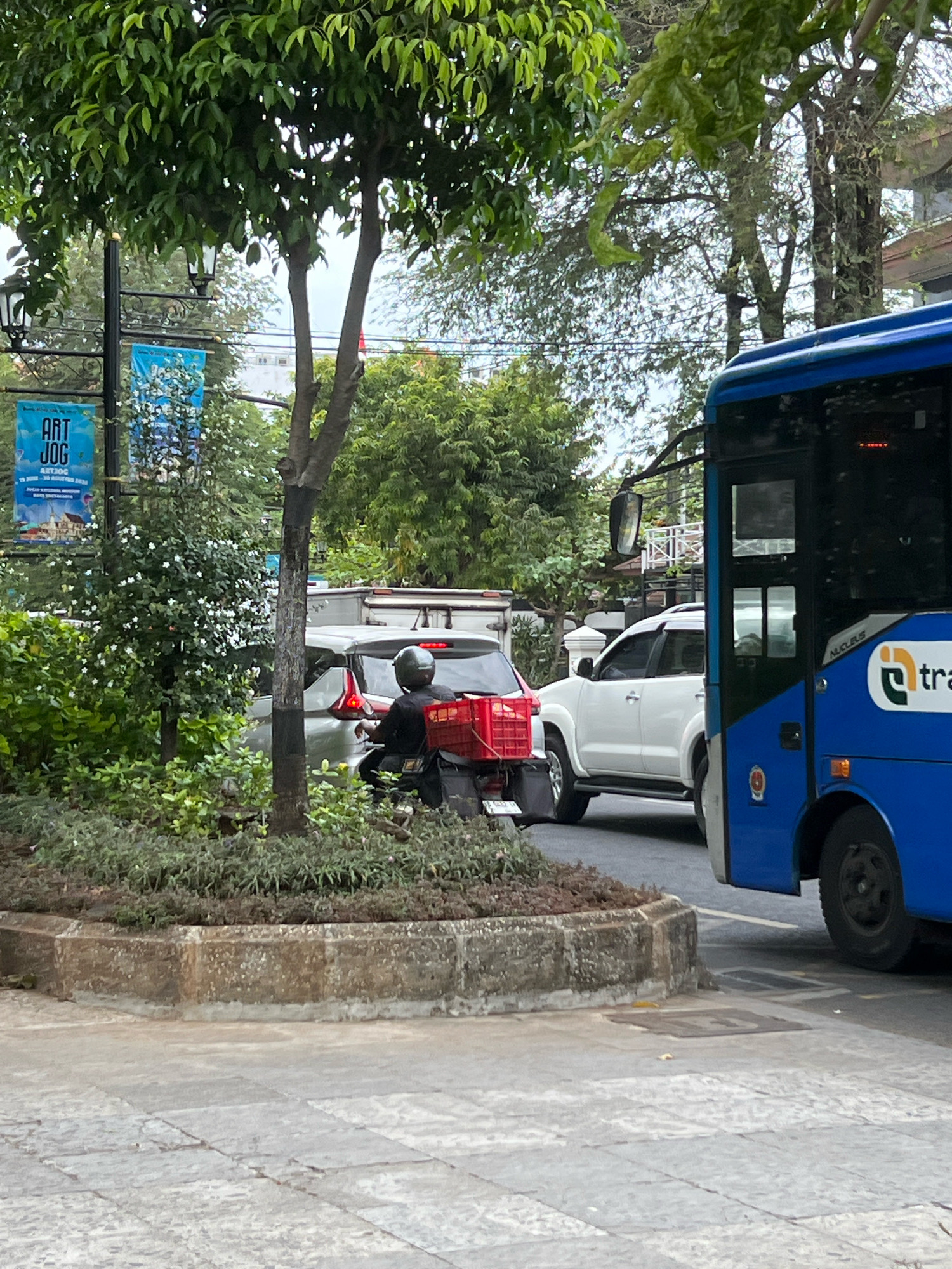 A medium shot captures a street scene in Yogyakarta, Indonesia, with a prominent blue bus on the right side. The bus has the text "TRANS" and a logo on its side. In the middle ground, traffic is visible, including a white SUV and a motorcycle. A person wearing a helmet is riding the motorcycle, which has a red crate attached to the back. Behind the motorcycle, a white car is parked. The environment is lush with trees and greenery, and a banner that reads "ART JOG" is visible on a pole to the left. The ground is paved with cobblestones, and a concrete curb is present in the foreground. The overall impression is a busy urban environment with a mix of vehicles and natural elements.
