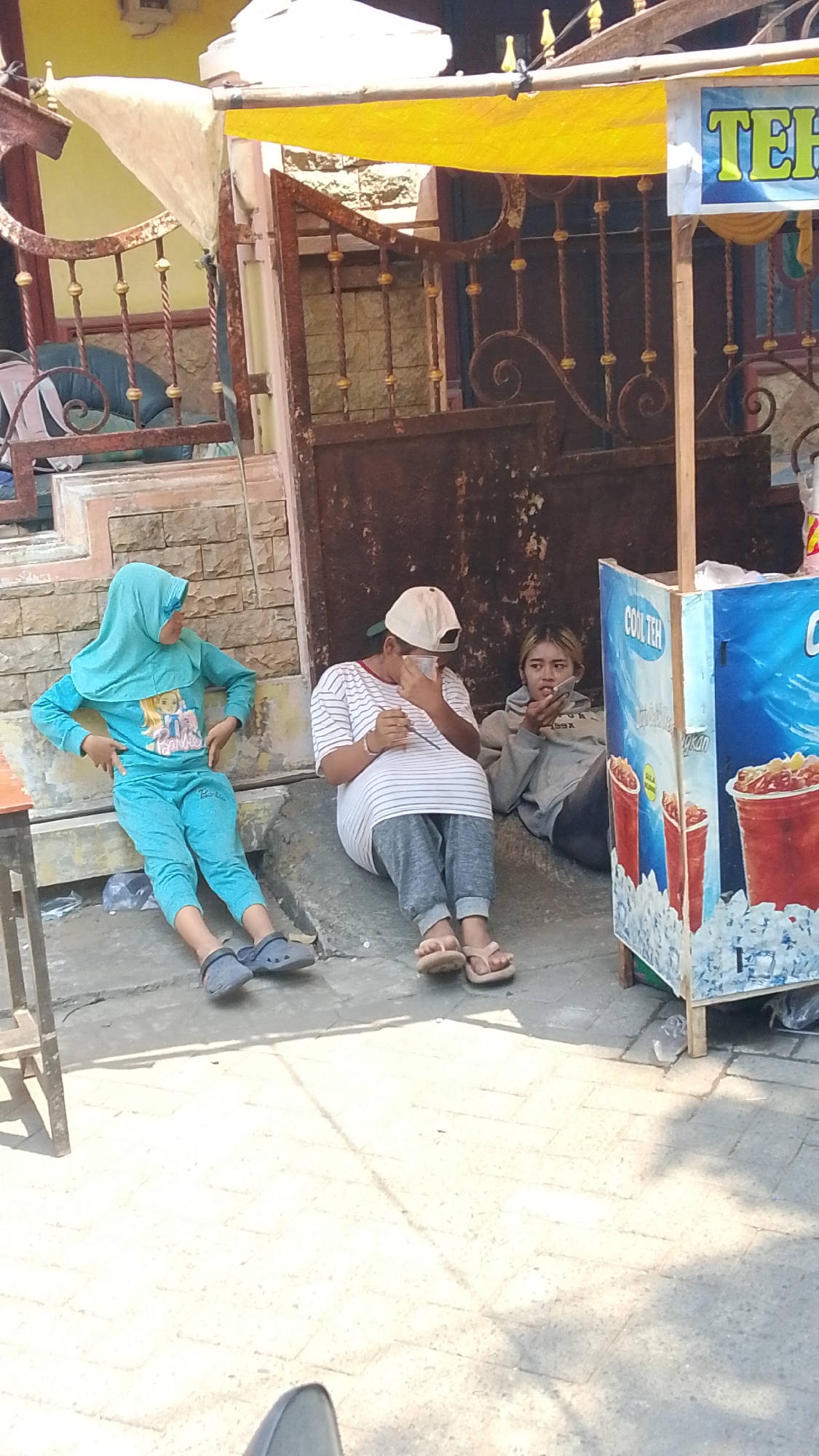 In Mancar, Indonesia, three individuals are seated near a street stall. On the left, a person wearing a turquoise hijab and matching tracksuit sits with their legs extended. In the middle, another individual in a white and striped long-sleeve shirt and grey pants is seated and appears to be drinking from a cup. To the right, a third person, wearing a grey hoodie, is also seated and holding a cup to their mouth. The street stall has a colorful advertisement depicting drinks with ice. The environment suggests a warm, sunny day with an outdoor setting.
