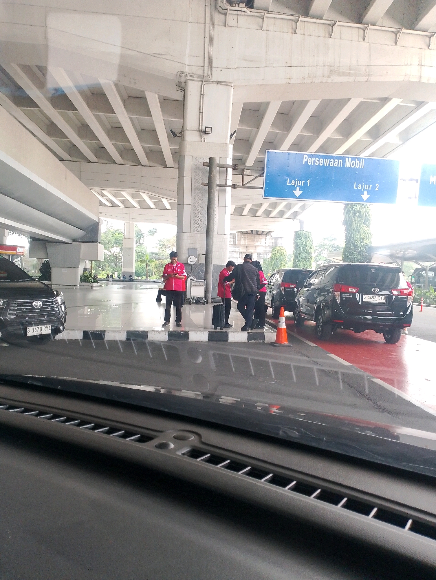 The image shows a view from inside a vehicle looking out at a car rental area in Tangerang, Indonesia. A sign overhead reads "Persewaan Mobil" which translates to "Car Rental." Below this, it indicates "Lajur 1" (Lane 1) and "Lajur 2" (Lane 2), both with downward-pointing arrows.
In the foreground, the dashboard of the vehicle is visible. Outside the vehicle, several black SUVs are parked. One SUV on the left has a license plate that reads "B 1670 RYA." Another SUV on the right has a license plate that reads "B 1492 DYU."
There are several people in the scene, appearing to be employees of the car rental company, identifiable by their bright red shirts. One person in a red shirt is standing and looking at a mobile phone. Another person in a red shirt is standing next to a suitcase. Two other individuals, one in a black jacket and another in a red shirt, are interacting near the vehicles.
The environment suggests an outdoor covered area, possibly a parking garage or an open-air terminal, with concrete pillars and an elevated structure above. The ground is a mix of pavement and a red-colored surface. Trees and greenery are visible in the background, indicating it's a location with some outdoor exposure. The lighting suggests it is daytime.