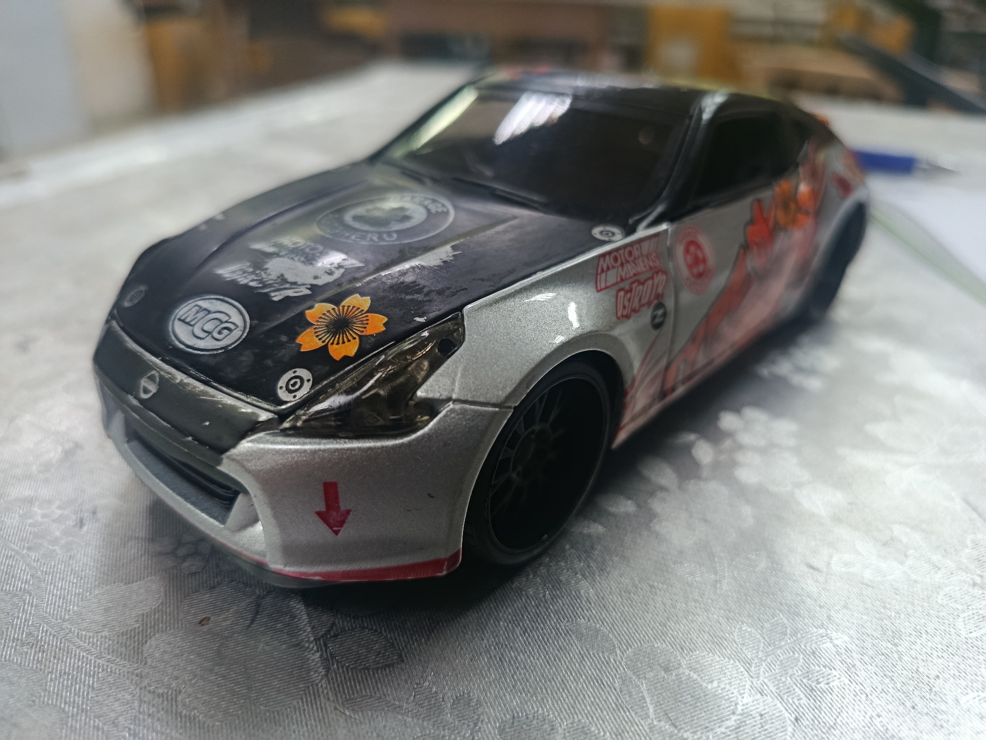 This is a close-up, eye-level, slightly angled, indoor shot of a miniature, customized toy car. The car is silver and black with various decals. It features a prominent orange cherry blossom design on the hood, a circular "MCG" logo, and other racing-themed stickers. The car is placed on a light-colored, textured surface, possibly a tablecloth or placemat. The background is blurred, showing indistinct shapes and colors that suggest an indoor environment. There is a blue pen visible in the upper right corner. The image does not depict the city of Sertolovo, Russia, or any of its features.