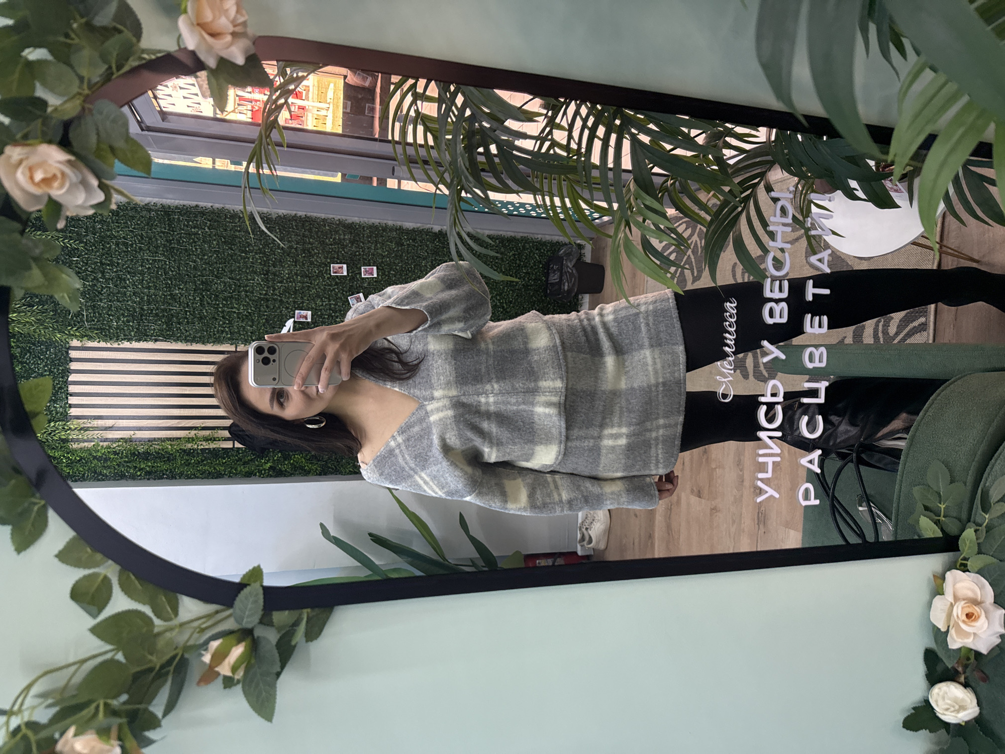 The image shows a person taking a mirror selfie. The person is wearing a grey and white plaid sweater and black leggings. The mirror is round and decorated with flowers and plants. The background of the mirror shows a store with clothes and a green wall. Text on the mirror reads, "Мелисса учись у весны расцветай" (Melissa, learn to bloom from spring). A location is suggested: Chaikovsky, Russia.
