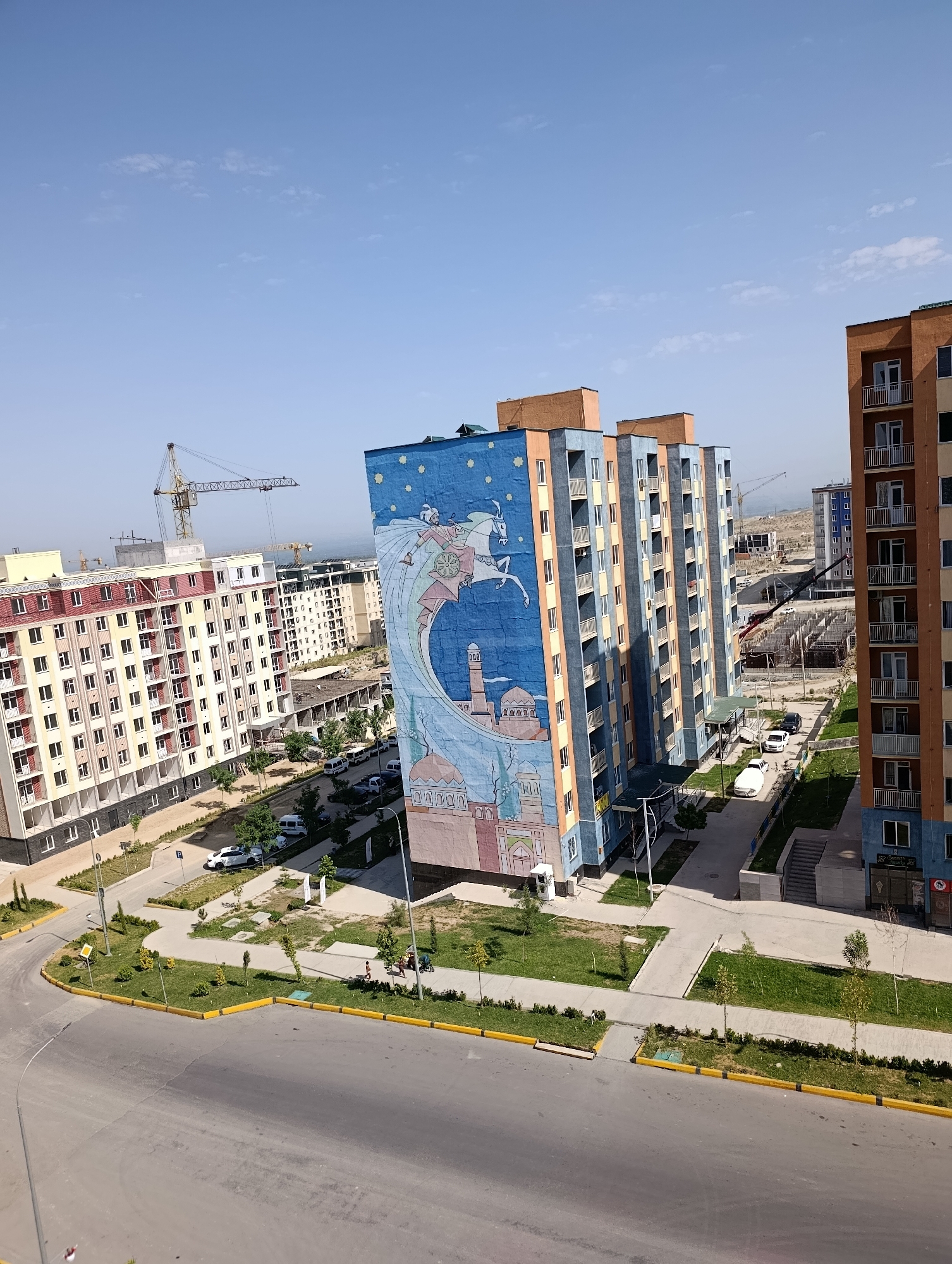 This is a high-angle, daylight exterior shot of a newly developing urban area in the city of Yangi Andijan, Uzbekistan. In the foreground, a wide asphalt road curves to the left, bordered by a yellow curb and green landscaping with small trees and shrubs. Several vehicles are visible on the road and in parking areas, including white sedans and a white van.
Dominating the midground are several modern, multi-story apartment buildings. The building on the far right is orange and white. Next to it is a cluster of three taller, narrow buildings, painted in alternating bands of blue and orange, with balconies. The leftmost of these blue and orange buildings features a large, vibrant mural covering its entire side. The mural depicts a historical or mythological scene with a rider on a horse within a crescent moon, a mosque with a minaret, and a domed structure, all set against a starry blue sky.
Further in the background, more apartment buildings are under construction, with cranes and scaffolding visible. The overall impression is one of rapid urban development. The sky is clear and blue with a few wispy clouds. The environment appears to be a residential district with amenities such as walkways and green spaces.