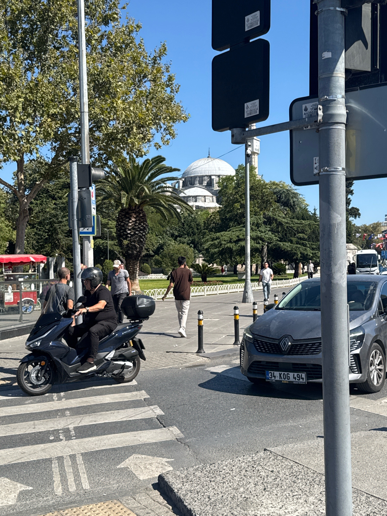 In Istanbul, Turkey, on a sunny day, a man rides a dark-colored scooter across a pedestrian crossing. The scooter has a black top box. Behind him, a dark gray Renault car waits at the intersection, its license plate reading "34 KOG 494". In the background, several people are walking on the sidewalk. Trees and a distinctive domed building, likely a mosque, are visible. Traffic signals and streetlights are present on a metal pole on the right side of the image. The environment appears to be a busy urban area with a clear blue sky.