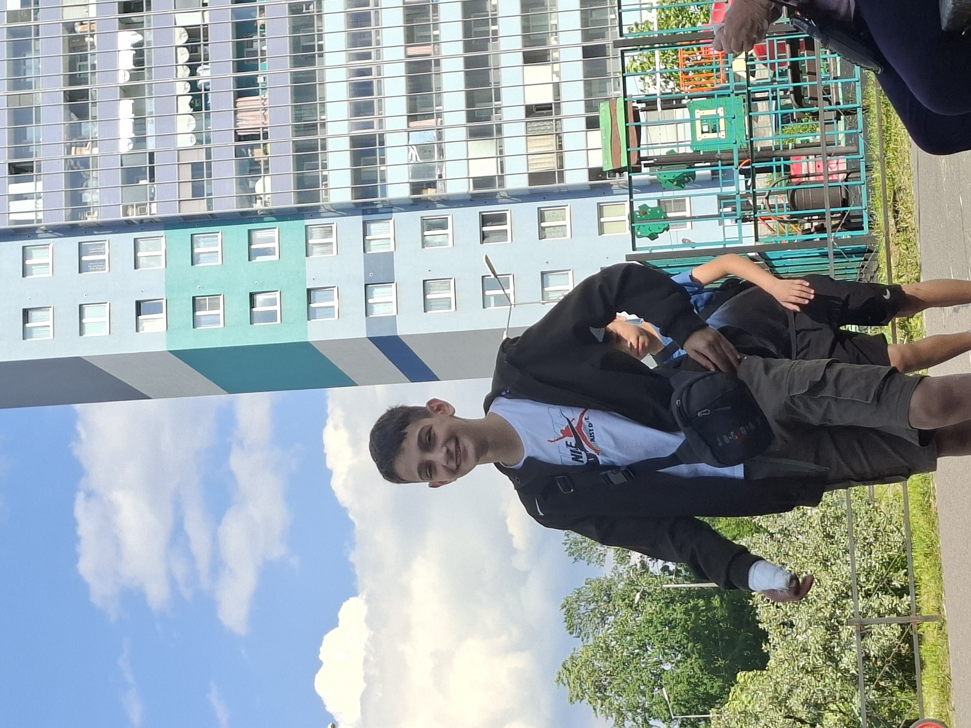 This image, set in Saint Petersburg, Russia, captures a bright, sunny daytime scene with several people in an urban environment. The primary subject is a young man, likely a teenager, positioned centrally and smiling warmly at the camera. He wears a black jacket over a white t-shirt featuring a "NIKE" logo and other text, along with dark cargo shorts. A black fanny pack, visible with the "JORDAN" brand, is worn across his chest. A distinct detail is a white bandage wrapped around his right hand.
In the background, a prominent multi-story building with alternating light blue and grey sections fills the upper left. Its facade is characterized by numerous windows, some reflecting the blue sky and clouds. Below this large structure, a smaller building section in varying shades of light blue and turquoise is visible, also dotted with windows.
To the right, another young man in a blue t-shirt and black shorts is partially visible, appearing to be lying down or resting near a colorful playground structure with green and red components. At the far right edge of the frame, the lower legs and feet of a third person are visible. Lush green trees and a patch of grass are present in the middle ground, indicating a park or recreational area adjacent to the residential buildings. The sky is clear blue with scattered white clouds, suggesting pleasant weather. The main subject's smile conveys a cheerful and relaxed demeanor.