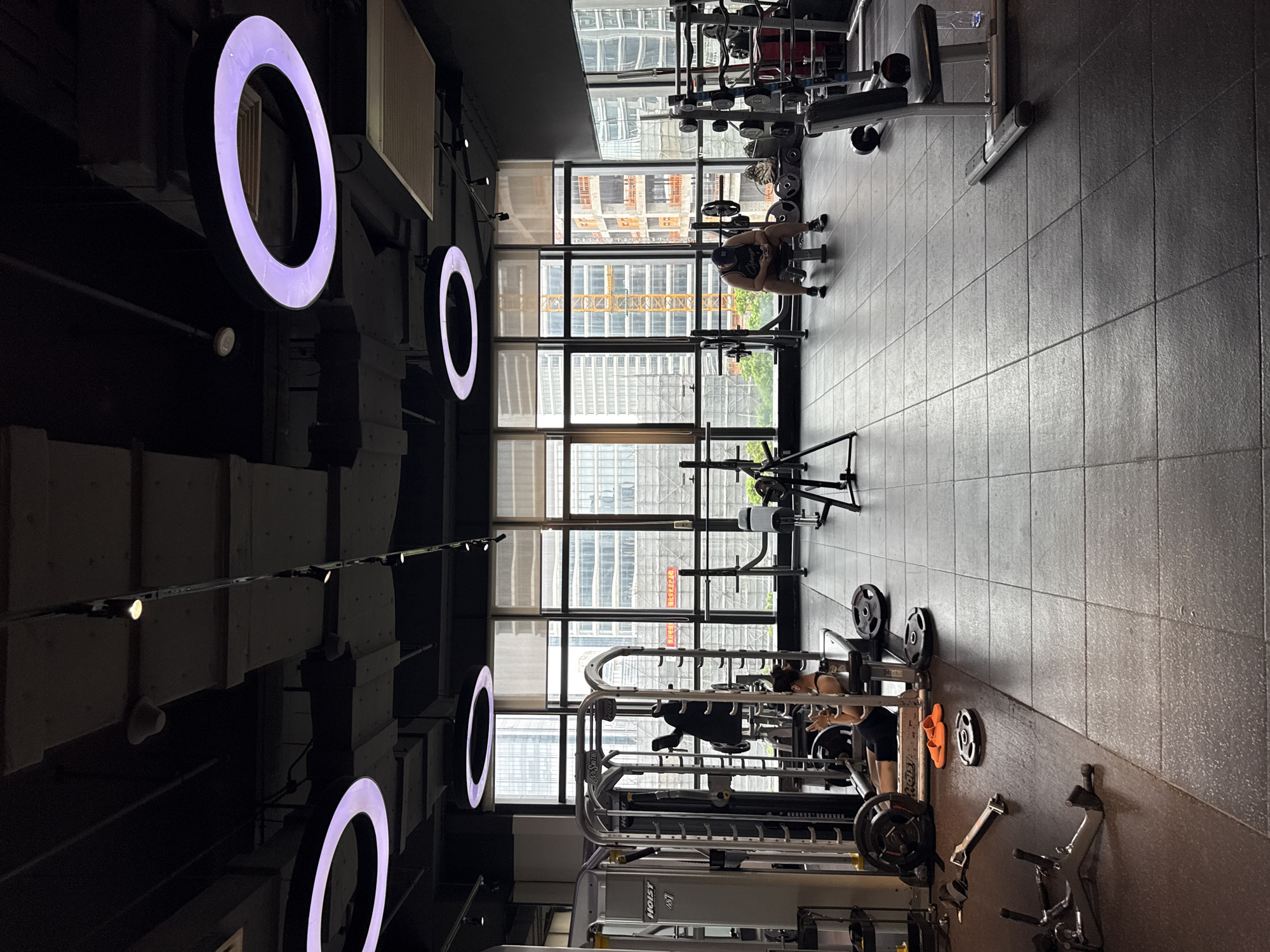 An interior shot of a gym in the city of Haizhu District, China, shows a spacious workout area with large windows overlooking an urban skyline. The gym features a variety of weightlifting equipment, including a multi-station gym machine on the left, weight racks with barbells and plates throughout the space, and benches. The floor is a dark, tiled surface. Two individuals are present: one person, appearing to be a woman, is seated at the multi-station gym, holding weights. Another person, appearing to be a man wearing a cap and a tank top, is sitting on a bench in the middle-right of the frame, looking down at a phone. Prominent circular light fixtures with purple illumination are suspended from the dark ceiling. The windows reveal numerous tall buildings, some under construction, indicating a dense urban environment.