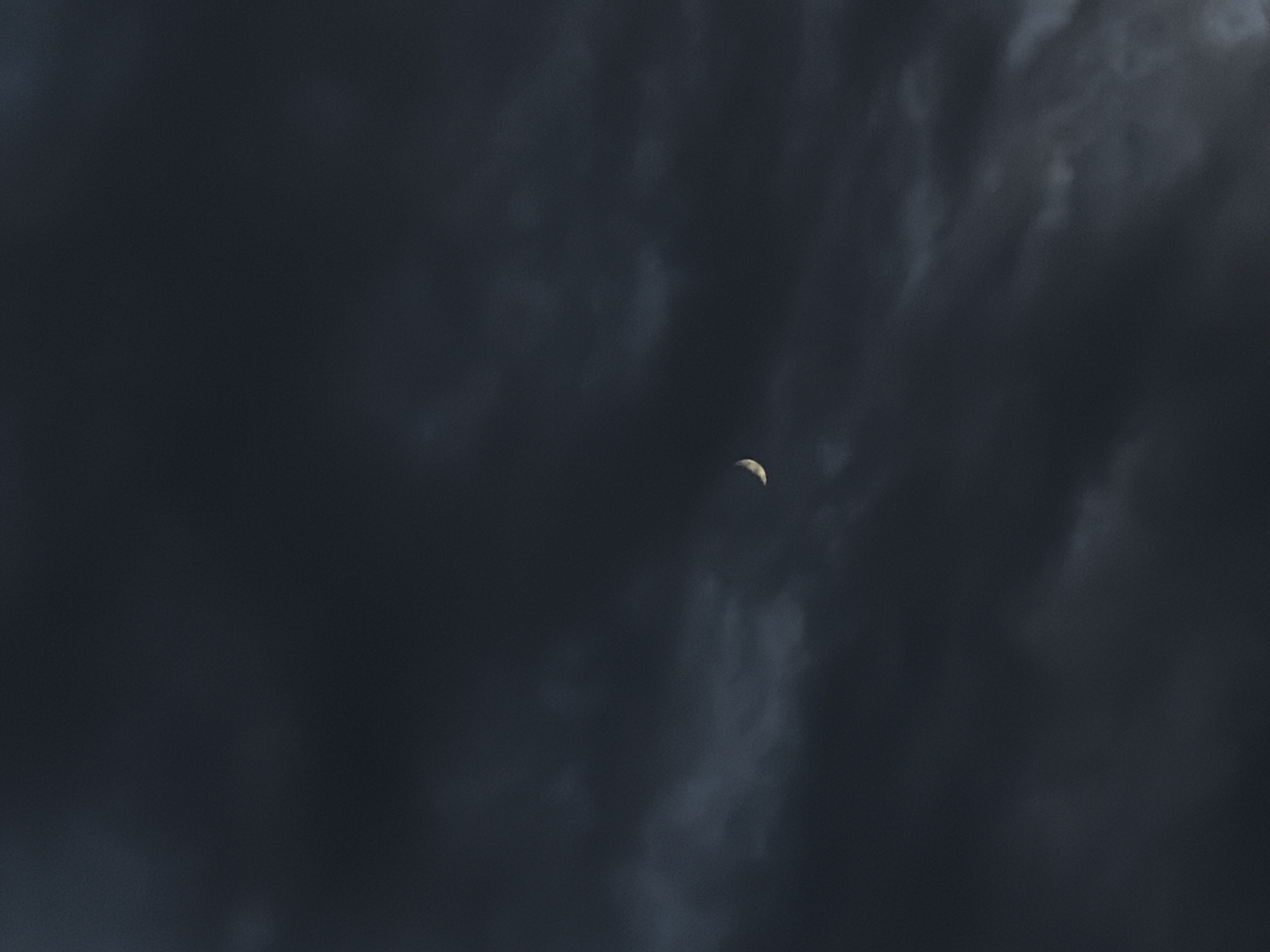 A crescent moon is visible in the dark, cloudy sky. The moon is a thin sliver of light against the deep blue-grey of the clouds. The clouds are thick and turbulent, suggesting an overcast or stormy atmosphere. The overall scene is dimly lit, with the moon being the only significant light source. The image appears to be a close-up shot of the sky.