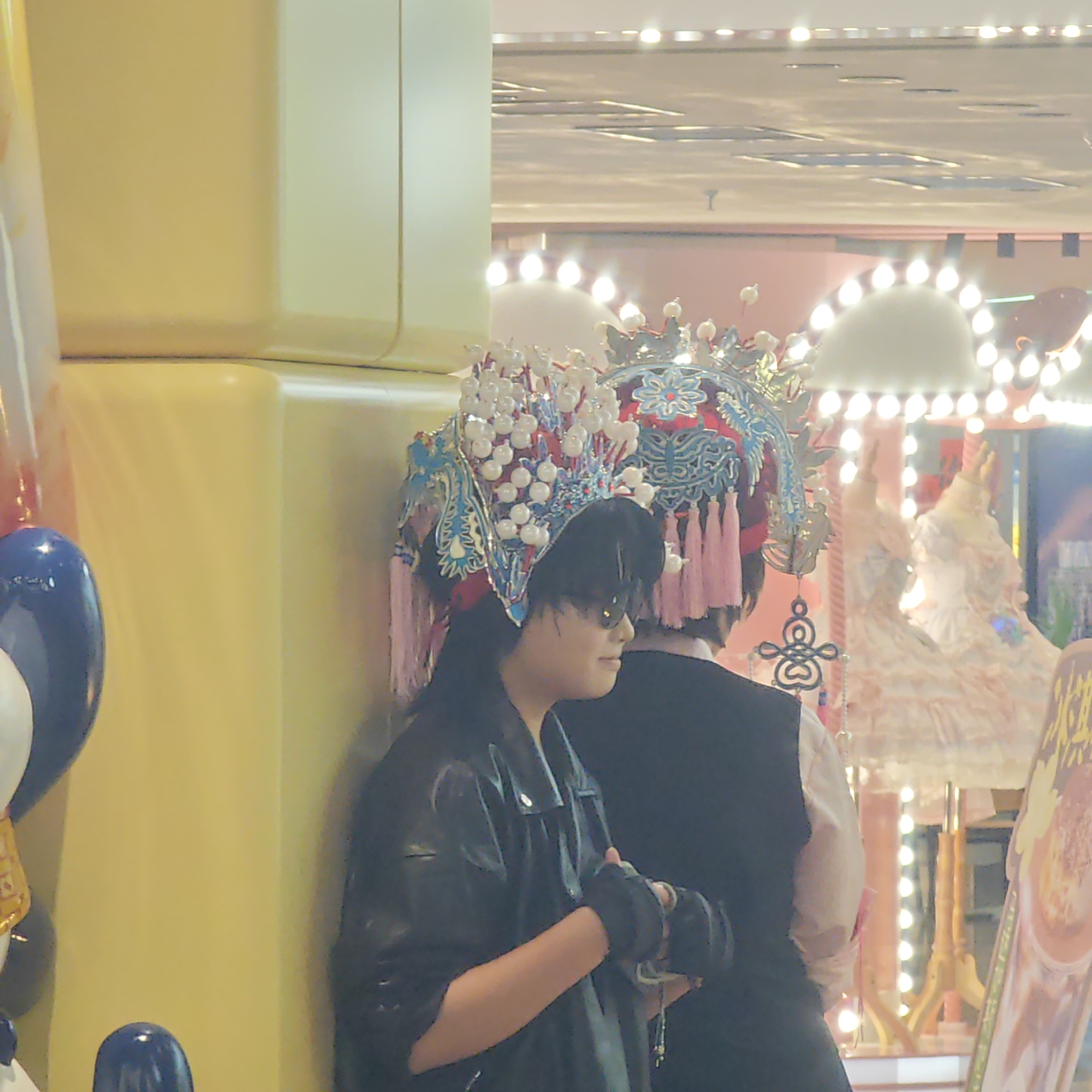 The image shows two individuals in a brightly lit indoor location, possibly a shopping mall or entertainment center. They are dressed in distinctive attire. The person on the left wears a black leather jacket, black gloves, and sunglasses, and a decorative headpiece with white pearl-like adornments and blue and red accents. The person on the right wears a vest and a similar ornate headpiece with pink tassels. Behind them, through a window or opening, there is a view of carousel-like decorations with bright lights. To the left of the individuals is a yellow pillar with balloons. The image suggests the scene might be a costume event or performance. Liangjiang, China is not explicitly confirmed by visual details.