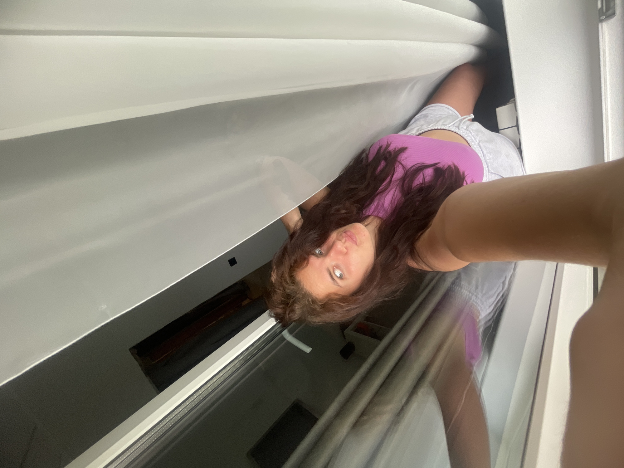 A person wearing a pink top and gray shorts is taking a selfie. The person has long brown hair and light-colored eyes. The photo is taken from a low angle, looking up. The background appears to be a window with white blinds. The location is unknown, but the city of Мосметаллоконструкция Онт, Russia, is mentioned in the query.