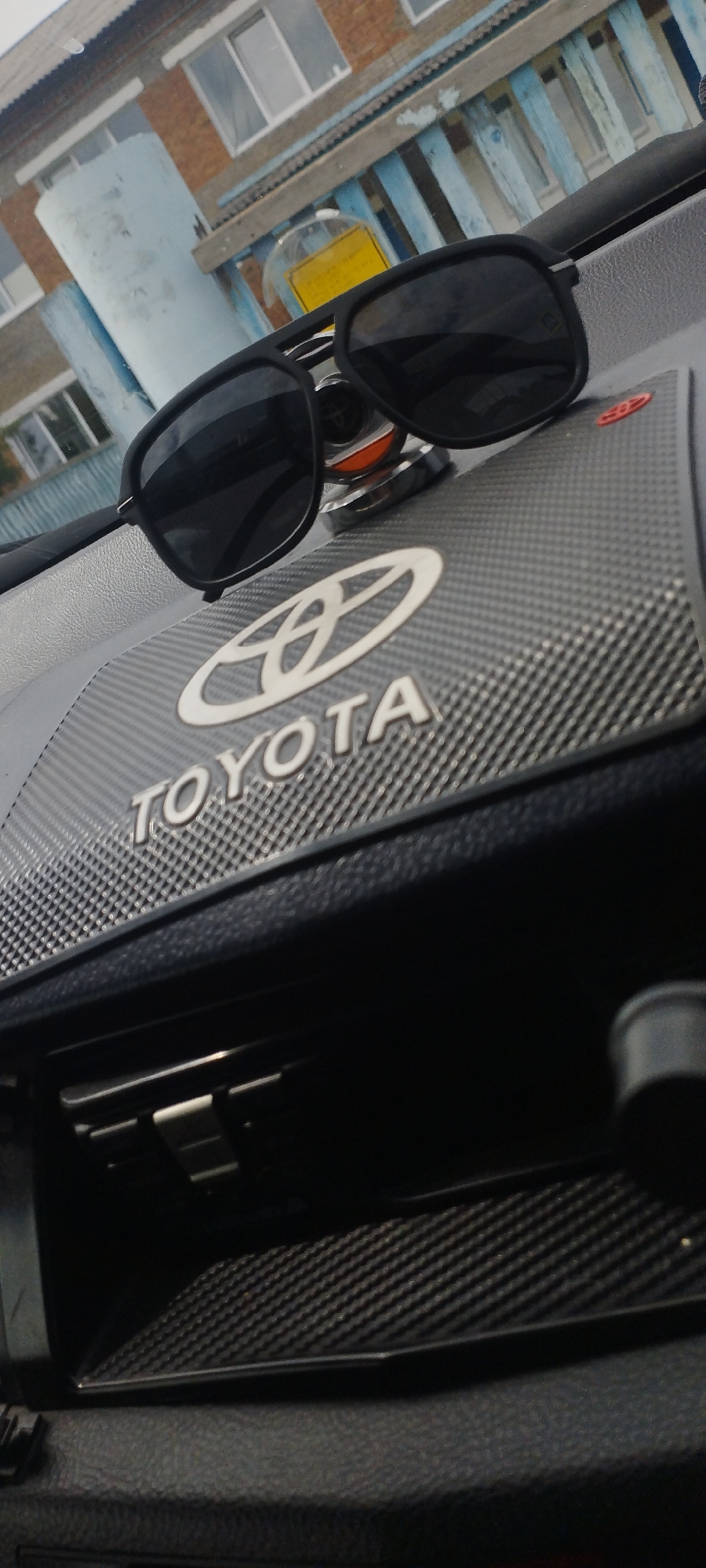 The image shows a pair of black sunglasses resting on a black textured dashboard with a silver Toyota logo. The dashboard is inside a vehicle. In the background, a light blue cylindrical object and a building with windows are visible through the windshield, suggesting an outdoor setting. The building has a textured facade and a blue railing. The environment appears to be urban or suburban, likely in a location with moderate weather conditions. The lighting suggests it is daytime. The car's interior, specifically the dashboard area, is the primary focus. No people are visible in the image. The location is identified as Pokosnoye, Russia.