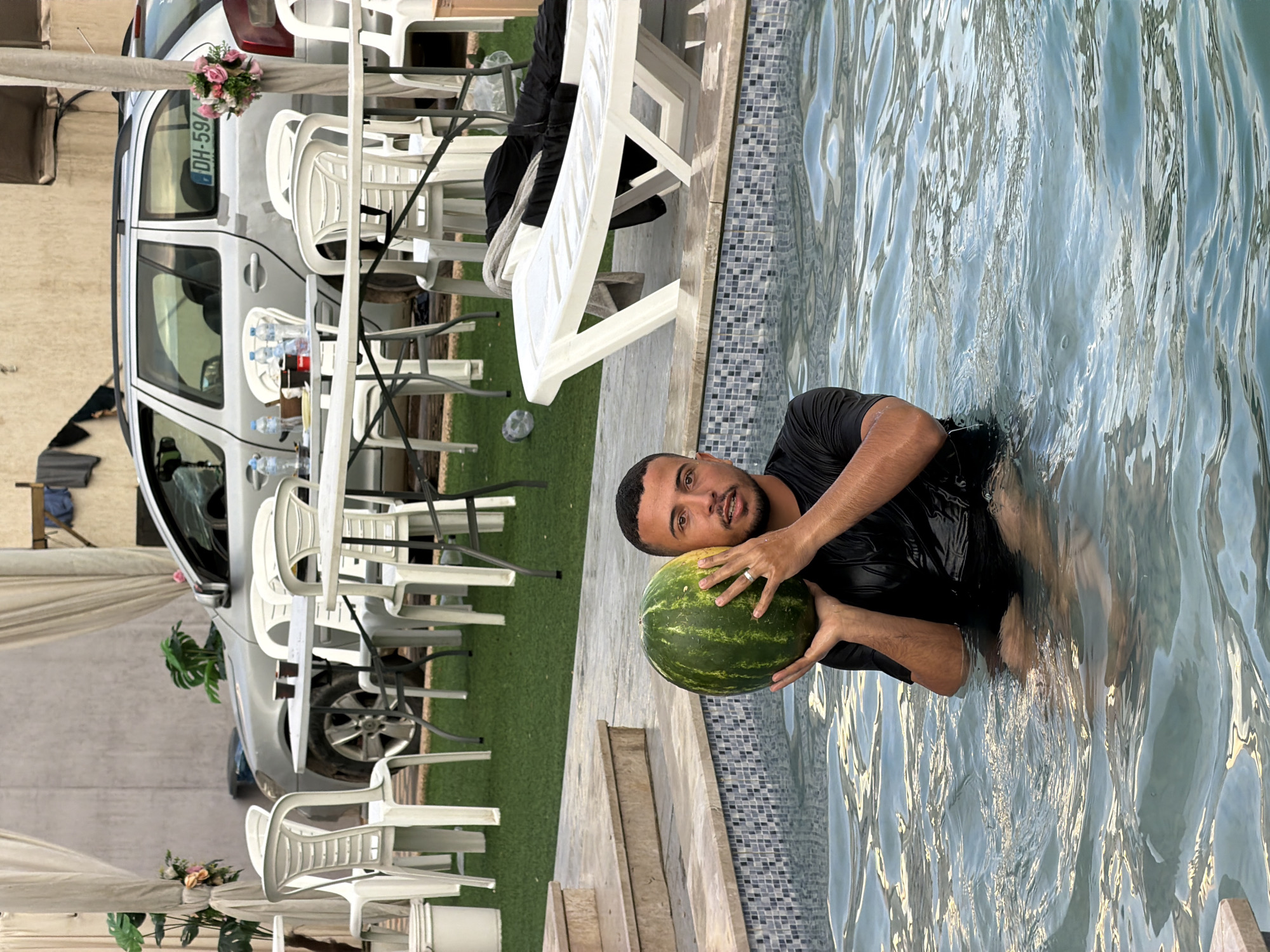 A man is holding a watermelon in a swimming pool. He is wearing a black shirt and shorts. The man is smiling and looking at the camera. The watermelon is green and has yellow stripes. The swimming pool is blue and has a tiled edge. In the background, there is a car, some chairs, and a table. There is also some grass and a canopy. The time of day appears to be daytime, and the weather seems to be pleasant. The man appears to be happy and relaxed. A license plate on the car reads "DH-597".