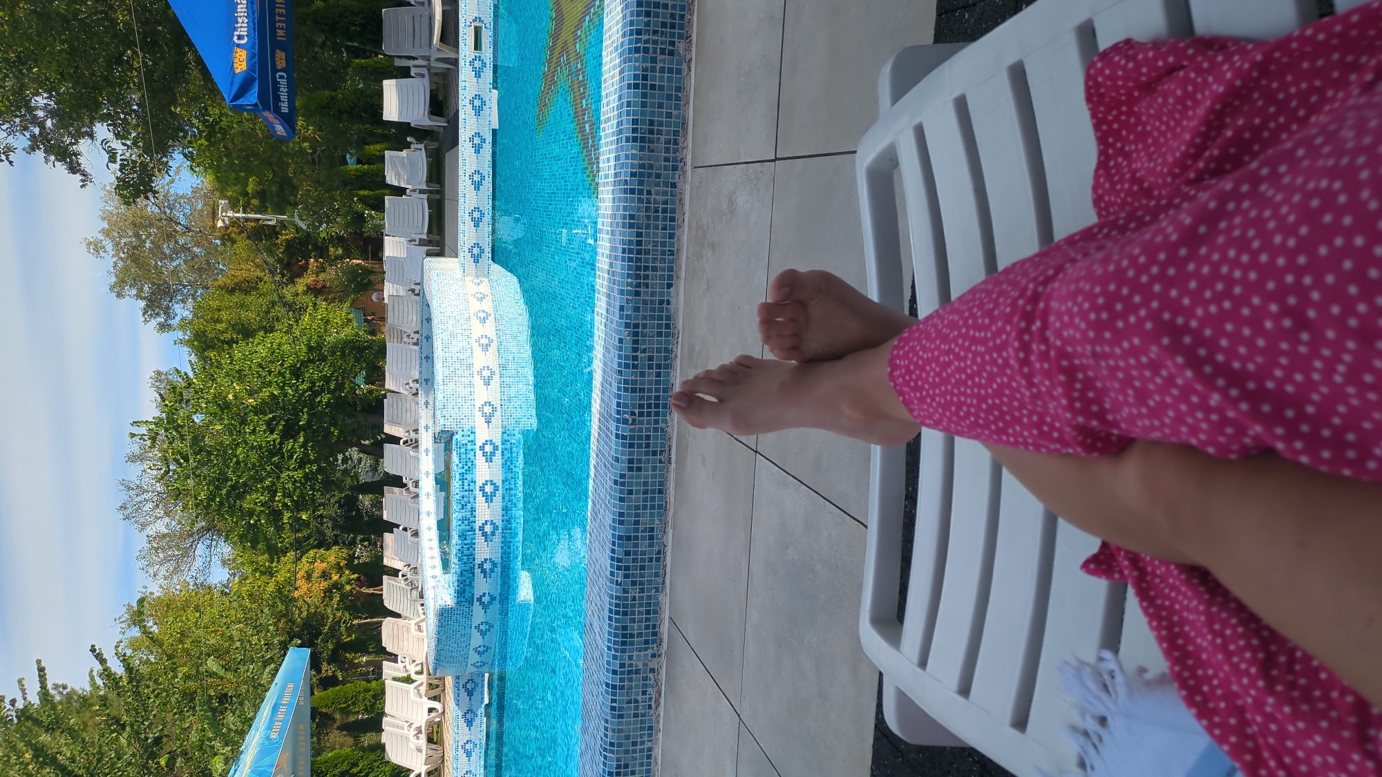 The image depicts a point-of-view shot of a person relaxing on a white lounge chair by a swimming pool. The person's bare feet, crossed at the ankles, are visible in the foreground, along with their legs covered in a pink garment with white polka dots. The swimming pool is bright blue and tiled around the edges with a mosaic pattern. In the background, a line of white lounge chairs is arranged by the pool, and beyond that, lush green trees and foliage are visible under a bright sky. Two blue umbrellas with white lettering are also present on either side of the scene. The overall setting suggests a sunny day at an outdoor pool, likely a resort or hotel, in Vadul lui Voda, Moldova, given the text visible on the umbrellas.