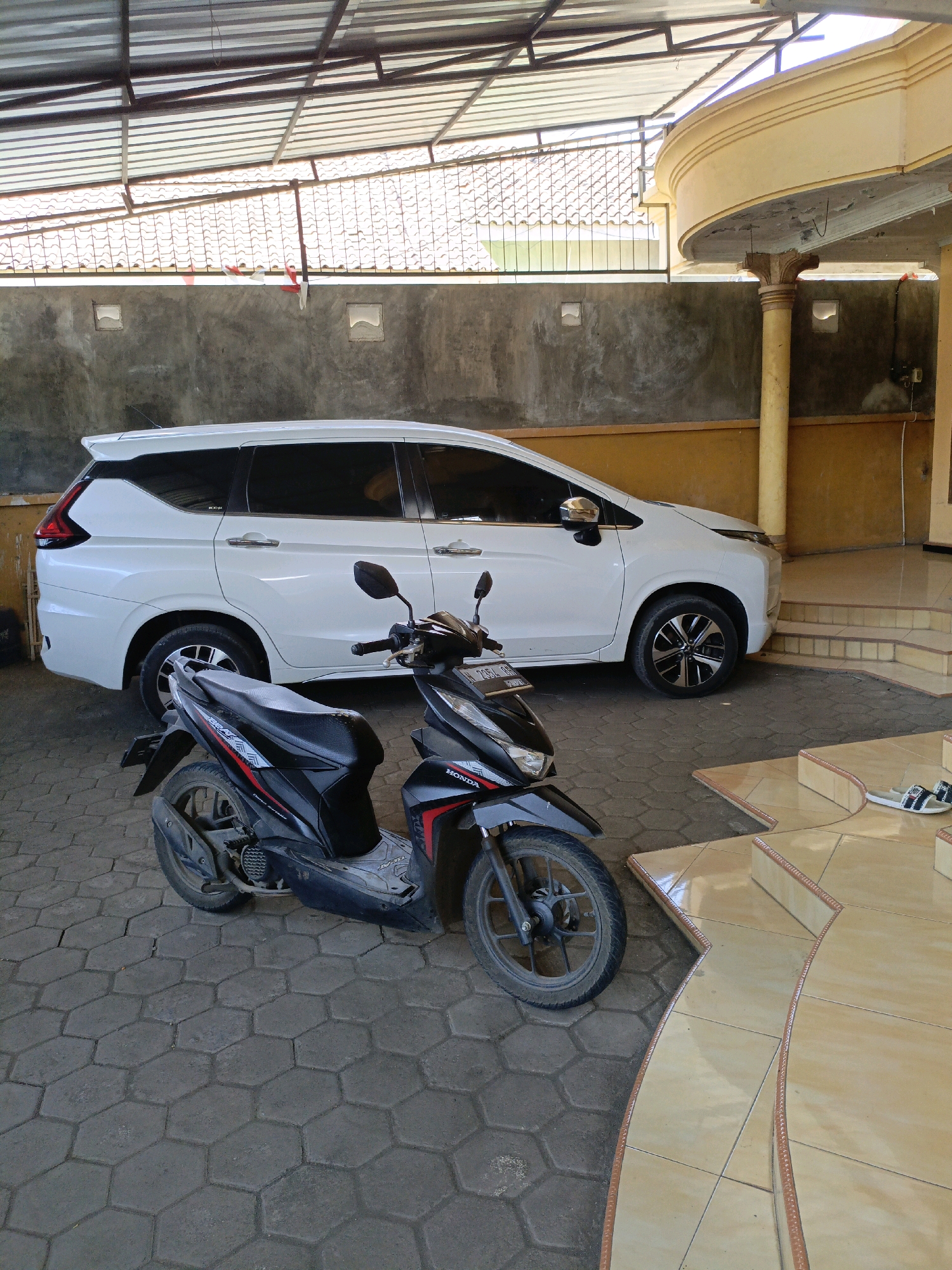 A black and white multi-passenger vehicle is parked next to a black and red motorcycle under a covered patio. The patio has a corrugated metal roof supported by metal beams. The wall behind the vehicles is concrete. The ground is paved with hexagonal concrete blocks. To the right of the vehicles are tiled stairs leading up to an entrance. A pair of white sandals with black stripes are placed on the top step. The scene is an outdoor area in Betek, Indonesia.