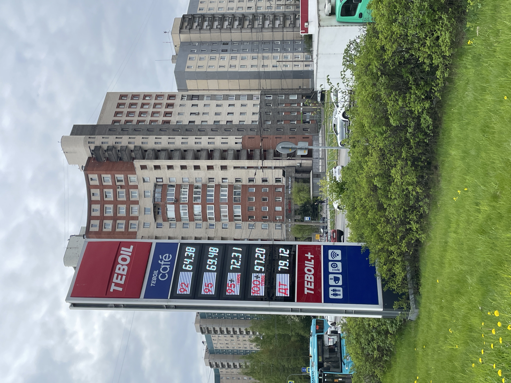 The image shows a Tehoil gas station price sign in Saint Petersburg, Russia. The sign displays the prices for different types of fuel, including 92 at 64.38, 95 at 69.48, 95+ at 72.31, 100 at 97.20, and Diesel at 79.12. The sign also includes logos for amenities such as Wi-Fi, car wash, a café, and restrooms. Behind the sign are tall apartment buildings with a mix of brick and concrete facades. A green bus is partially visible at the bottom of the frame, and there is a street with cars and a restaurant sign in the background. The surrounding environment includes greenery, with trees and grass visible. The sky is overcast.
