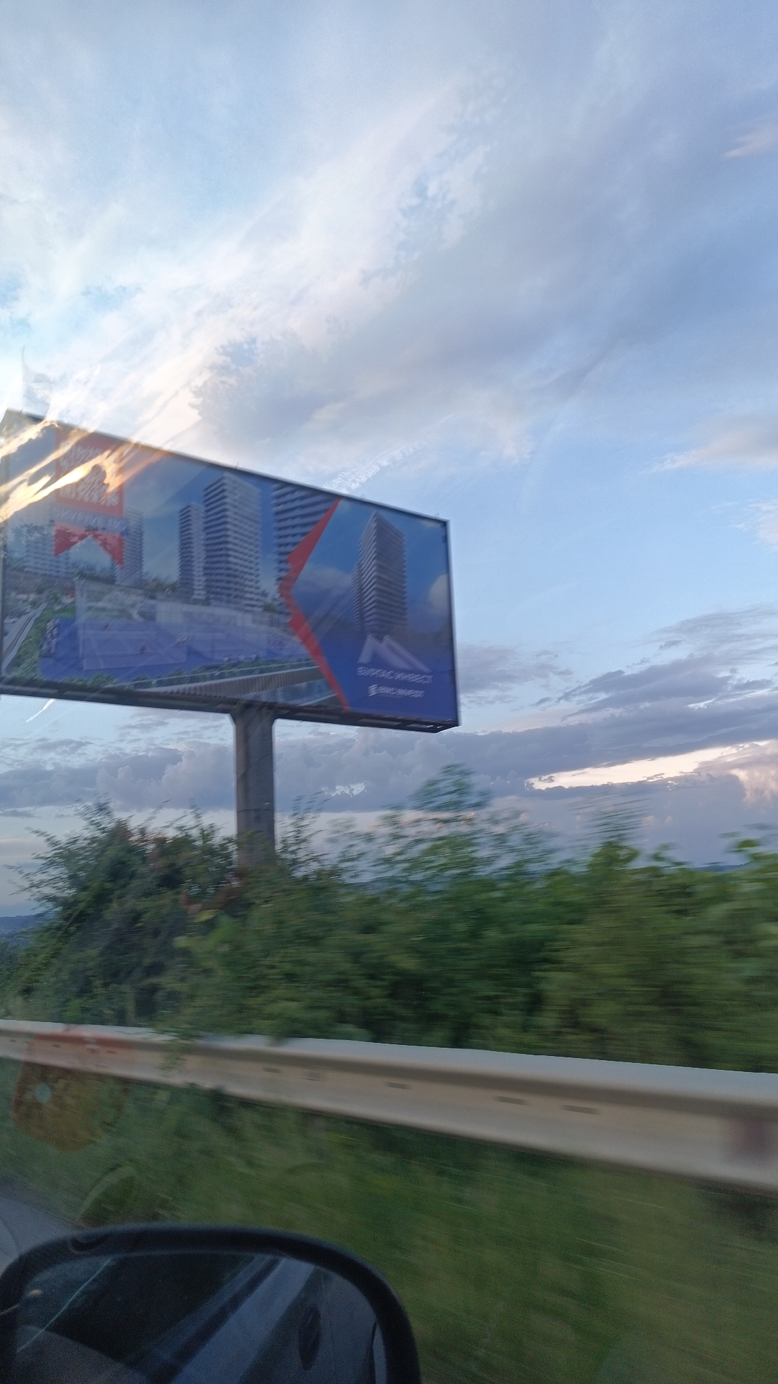 The image shows a billboard on a roadside, likely taken from a moving vehicle. The billboard advertises "Burgas Invest", showcasing images of modern buildings and a swimming pool. Below the billboard is green vegetation. A white guardrail is visible in the foreground, along with the side mirror of the vehicle. The sky is filled with clouds, suggesting either early morning or late afternoon. There are no people visible in the image. The location is likely in the city of Burgas, Bulgaria, as suggested by the billboard's branding.
