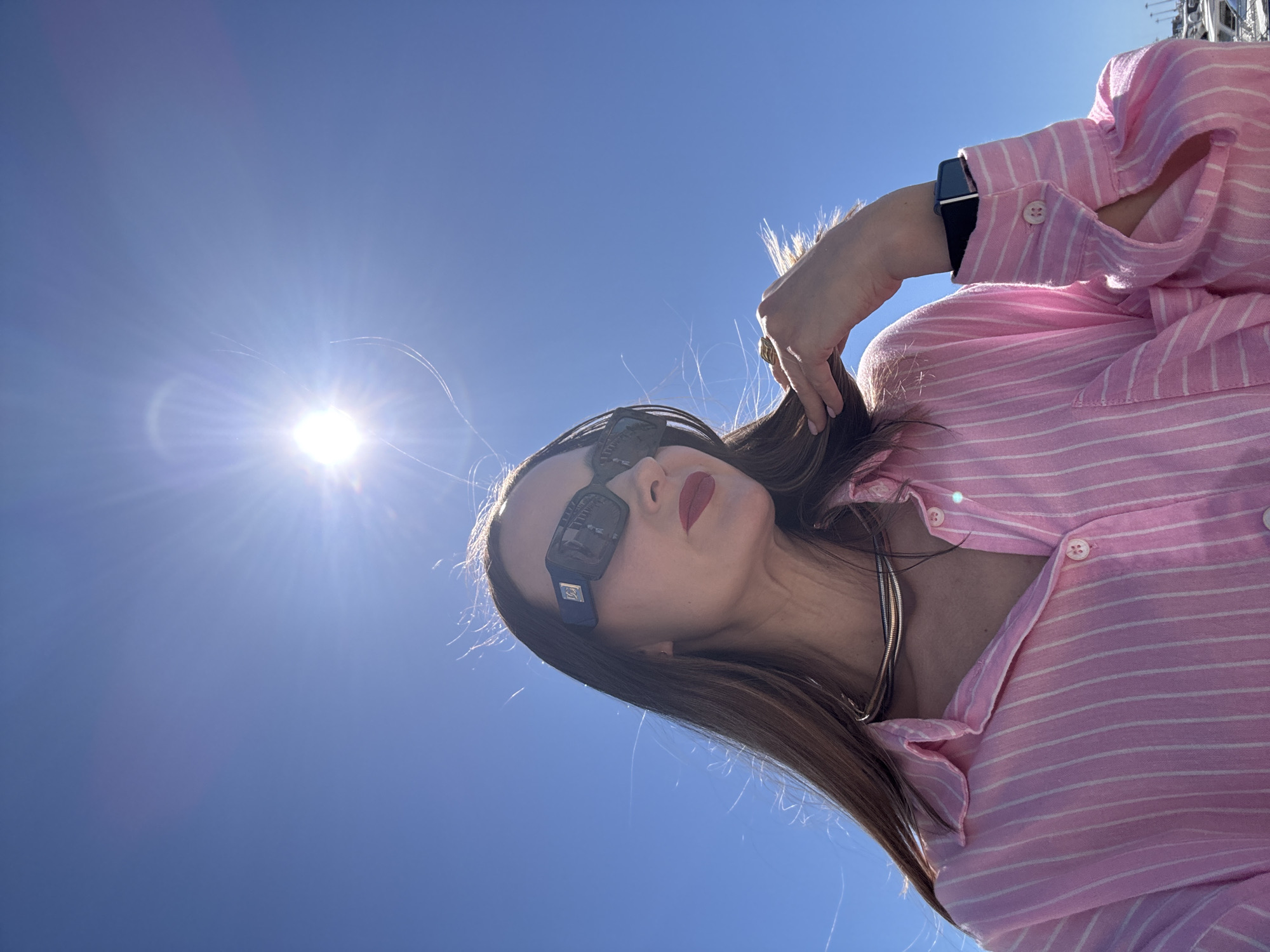 An outdoor, low-angle shot captures a person looking upwards against a clear, vibrant blue sky in Sochi, Russia. The bright sun, positioned in the upper left, casts strong light and creates a sunburst effect.
The main subject is a woman, angled in the lower right portion of the frame. She wears a pink and white striped long-sleeved shirt, which is partially unbuttoned to reveal a gold or silver-toned snake chain necklace. Her long, dark hair flows down her back and shoulder, and her left hand is raised, gently touching her hair. She wears dark, rectangular sunglasses with a visible small blue emblem on the side arm, featuring a white intertwined "CC" logo, indicative of Chanel. Her lips are painted with red lipstick. A black smartwatch or fitness tracker is visible on her left wrist, and a simple ring is on a finger of her left hand.
The activity appears to be posing for a photo under direct sunlight. In the top right background, barely visible against the blue sky, are faint outlines of what appear to be communication towers or antennas. The bright sun and cloudless sky indicate a clear, sunny day, likely midday or early afternoon. The overall impression is one of a relaxed moment captured in good weather.