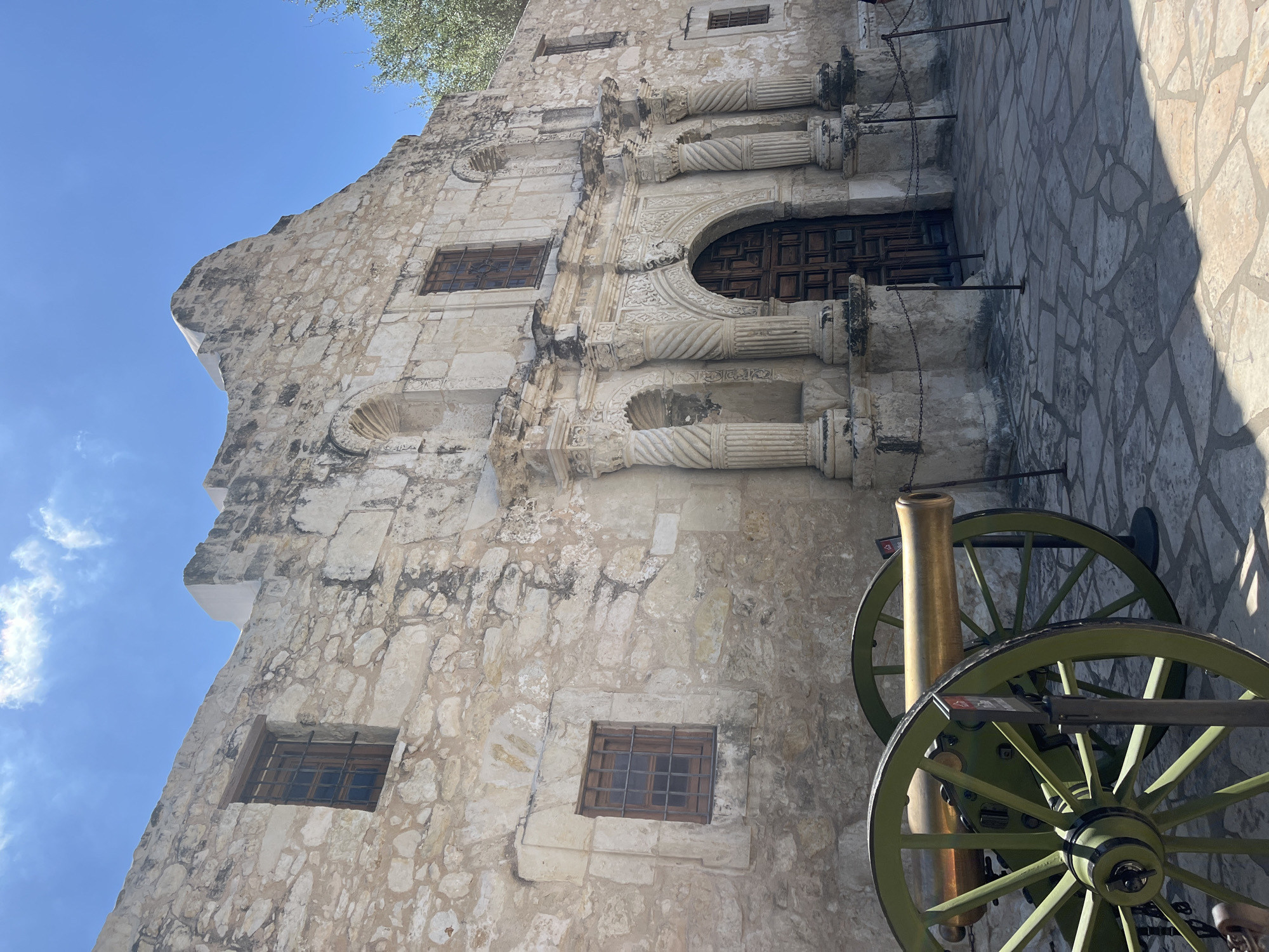 The image shows a ground-level view of the Alamo, located in San Antonio, Texas. The Alamo is a historic mission and fortress complex. The primary subject is the Alamo's facade, constructed of light-colored stone blocks. It features architectural details like columns, arches, and windows with dark frames. A large, ornate wooden door is visible at the building's center. A green and gold cannon stands in the foreground. The environment is a sunny day with a clear blue sky. The ground is paved with stone tiles. The image is taken at an angle, looking upwards at the building.