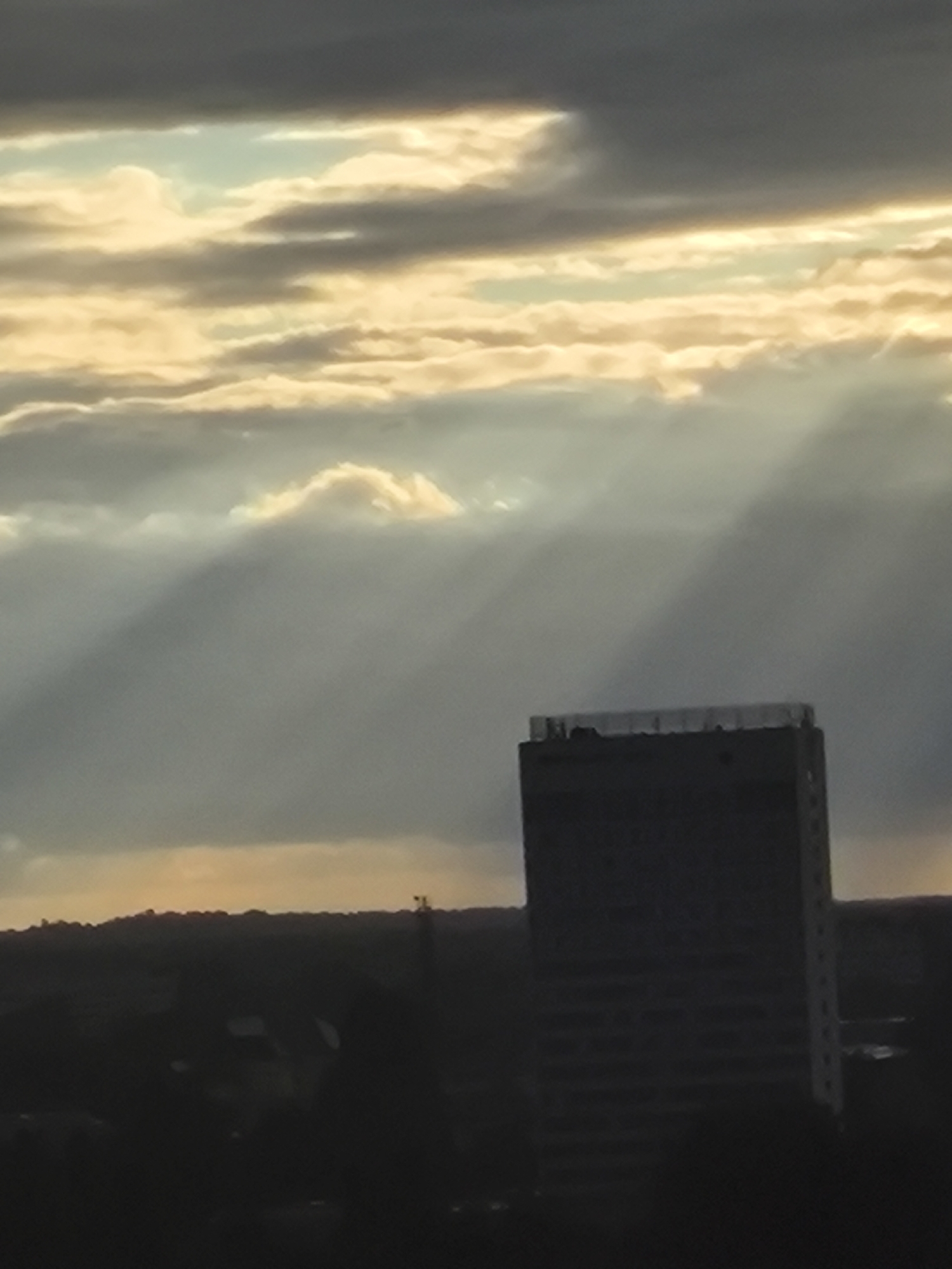 A tall, dark, rectangular building with a flat roof is silhouetted against a cloudy sky. Sunbeams are shining through gaps in the clouds, casting shadows that stretch across the scene. The building appears to be located in an urban environment, with a dark, indistinct landscape visible in the foreground. The sky is a mix of dark gray and bright yellow clouds. The image captures a moment of dramatic lighting, possibly during sunrise or sunset.