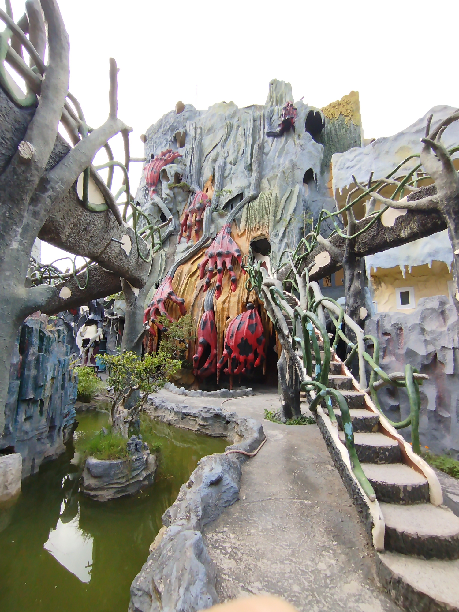 The image depicts the exterior of the Crazy House (Hang Nga Guesthouse) in Da Lat, Vietnam. A pathway made of concrete slabs and stone stairs leads towards a building resembling a cave or grotto structure. The building's facade is gray with organic, cave-like openings and red, black-spotted protrusions, possibly resembling fungi or abstract figures, jutting from the walls. A small, green-tinged pond with an island of greenery sits adjacent to the pathway. Man-made tree-like structures with painted branches frame the scene. The sky is overcast and white.