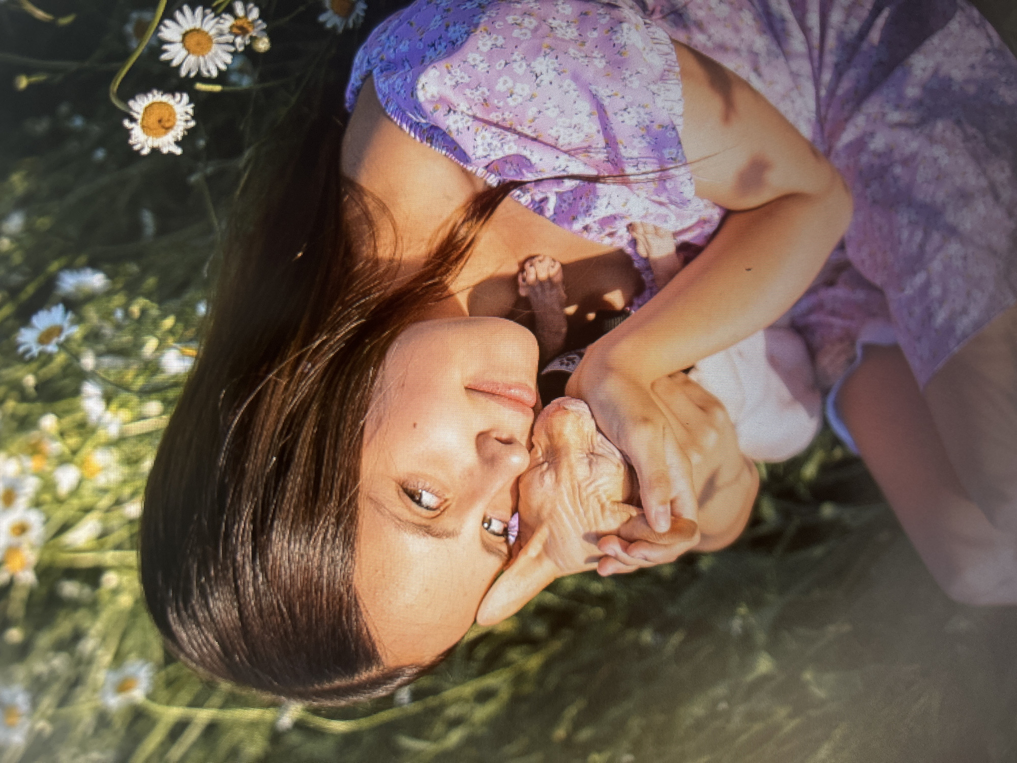 A young woman with long, dark hair and brown eyes is lying in a field of daisies, holding a Sphynx cat. She is wearing a purple floral dress. The woman is looking at the camera with a slight smile. The cat is nestled in her arms, and it appears to be asleep. The background is a blur of green grass and white daisies. This scene is not specifically identifiable as being in Kiselyovsk, Russia, or any other specific location.