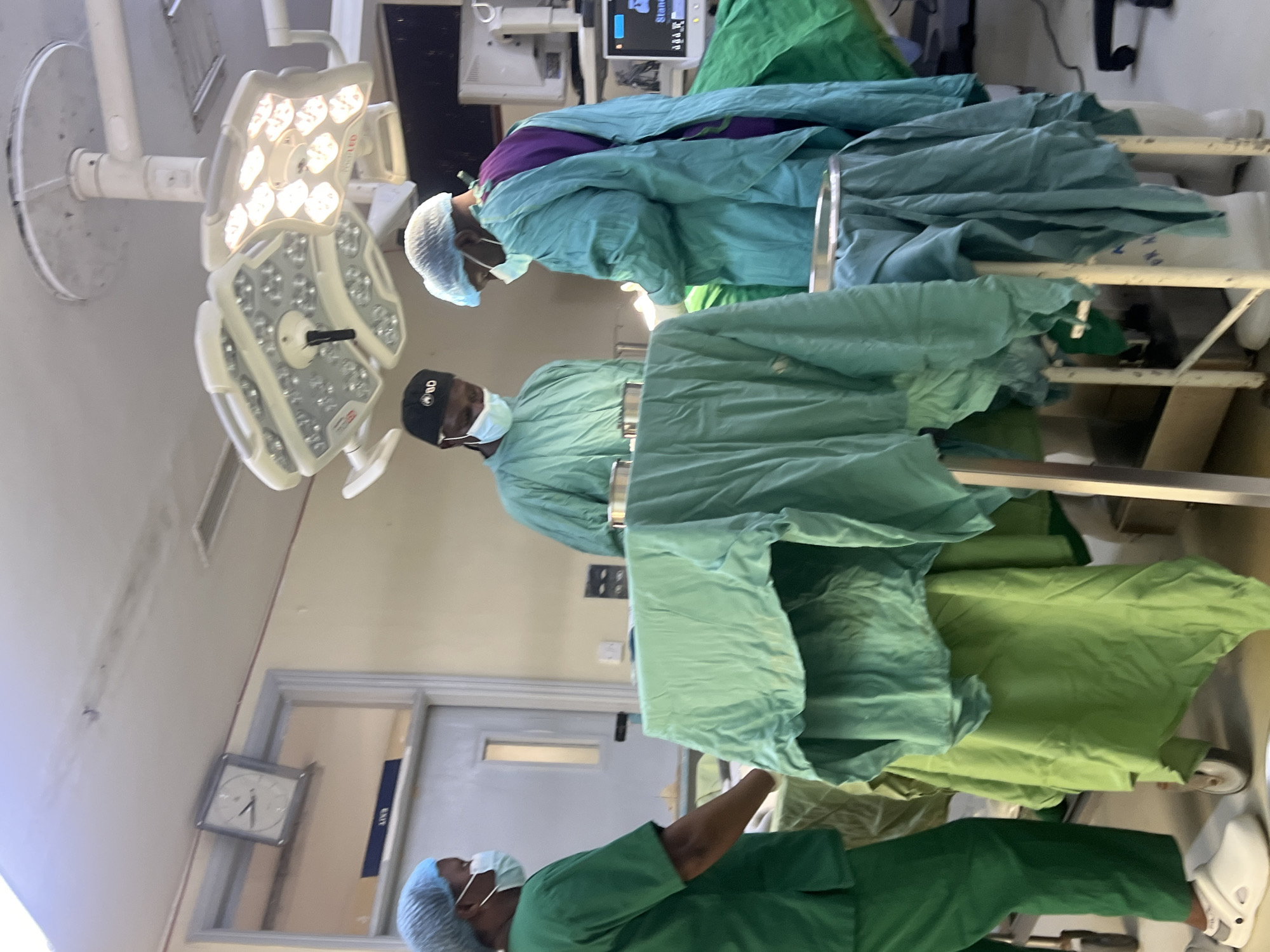 Three people wearing surgical scrubs and caps are visible in what appears to be an operating room. Two individuals are standing next to a surgical table draped in green cloth. A surgical light fixture is mounted on the ceiling. A clock is visible on the wall, and medical equipment can be seen in the background. It is not possible to confirm the specific location in Nairobi, Kenya, from this image alone.