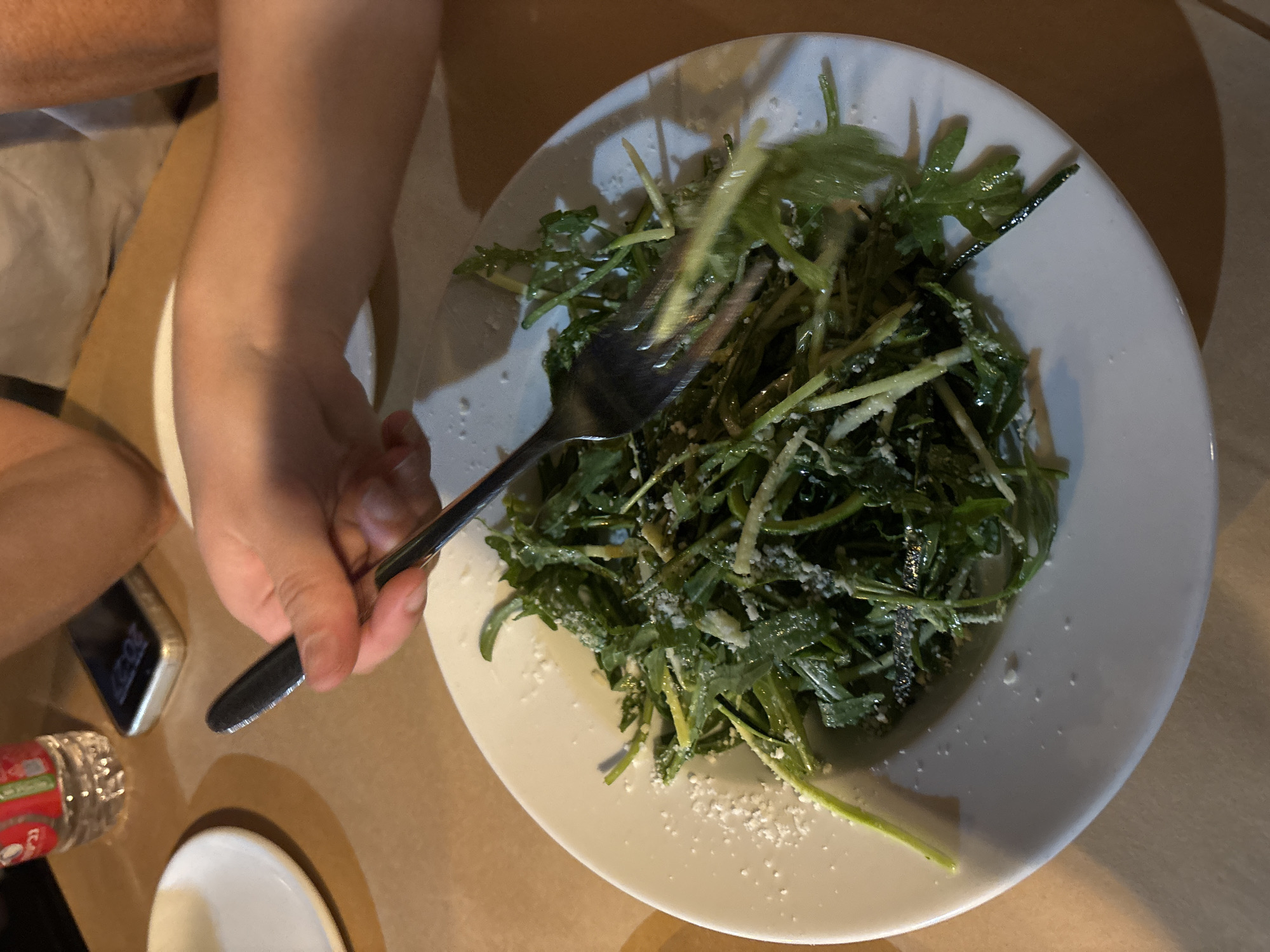 An overhead, slightly angled shot captures a dining scene, likely in a restaurant or cafe. The central subject is a white, round plate containing a vibrant green salad, possibly arugula, generously coated with what appears to be grated cheese or a creamy dressing, along with some thinly sliced green vegetables. A silver fork is held by a fair-skinned hand, entering the frame from the left, poised within the salad.
The table surface is a light brown, possibly wood or a placemat. The lighting is warm and subdued, creating soft shadows and suggesting an evening setting. This is corroborated by a partially visible smartphone on the lower left, which displays the time "20:27" (8:27 PM) on its dark screen. Next to the phone, a small, white empty dish and a plastic bottle with an unreadable red label are also visible. A person's arm or leg is partially seen on the far left. The image depicts the act of eating a salad during an evening meal.