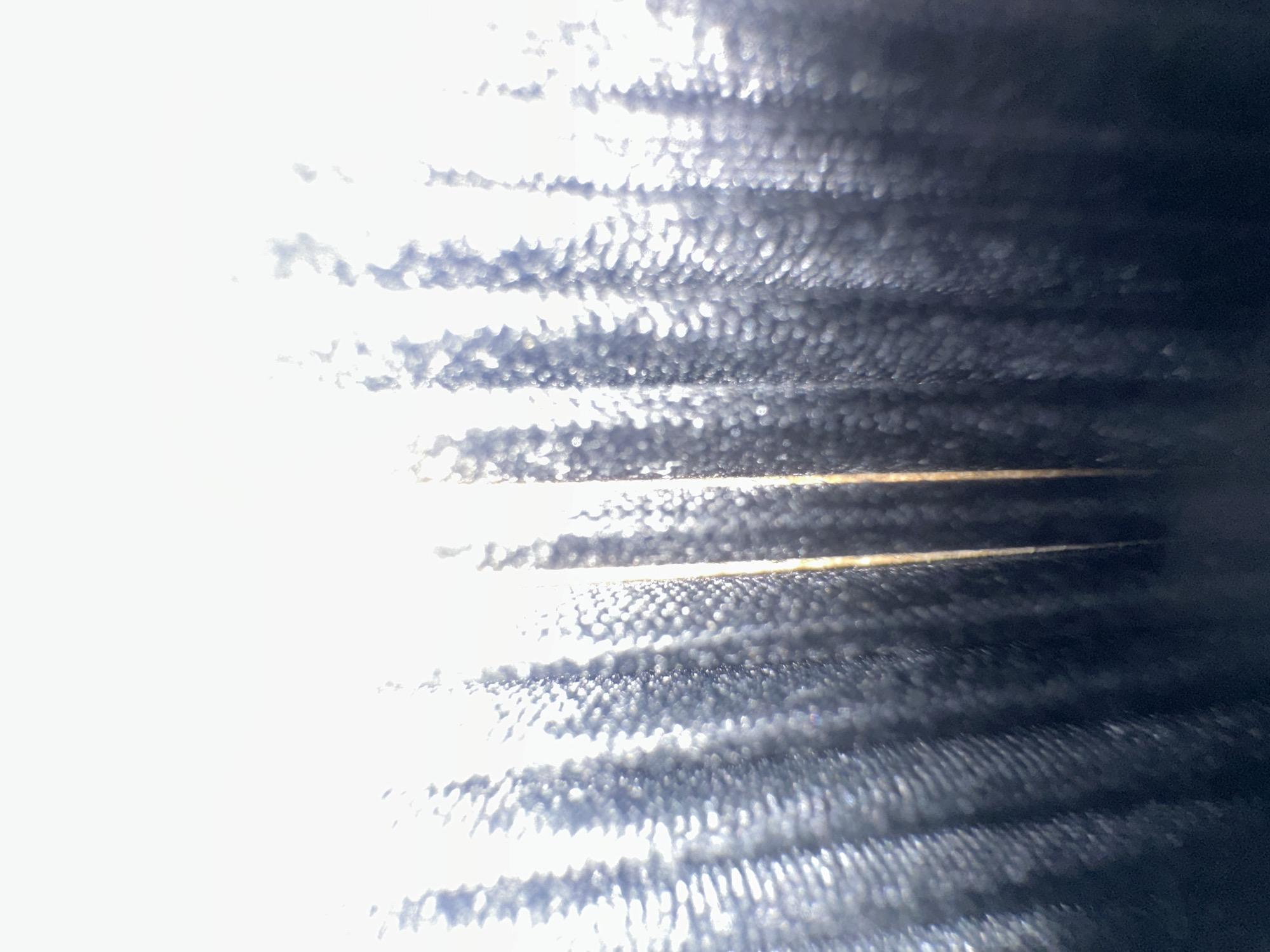 The image is a close-up, abstract shot of what appears to be the fins of an air conditioning unit or a similar heat exchanger. The subjects are the metallic fins, arranged horizontally and running from the foreground towards the background, which is mostly dark and out of focus. There are no people visible.
The activity is not apparent from the still image; it is a static view of an object. The setting appears to be indoors or close to a building, given the nature of the object depicted.
Notable details include the interplay of light and shadow on the fins. The left side of the image is brightly lit, creating a blown-out effect with strong highlights that obscure detail. As the eye moves to the right, the fins become more defined, revealing a textured surface. There are horizontal bands of lighter and darker tones, suggesting the presence of multiple layers of fins. Some of the fins have a slightly golden or coppery hue, while others appear silvery or dark gray. The texture of the fins is finely ridged or corrugated, and the bright light catches these ridges, creating a sparkling, almost crystalline appearance in some areas. The darkness on the far right suggests the fins are part of a larger, possibly enclosed apparatus. There is no indication of the time of day, weather, or specific emotions, other than perhaps a sense of industrial or mechanical detail. No text is visible in the image. The location context of Belgrade, Serbia, does not provide specific visual cues within the image itself.