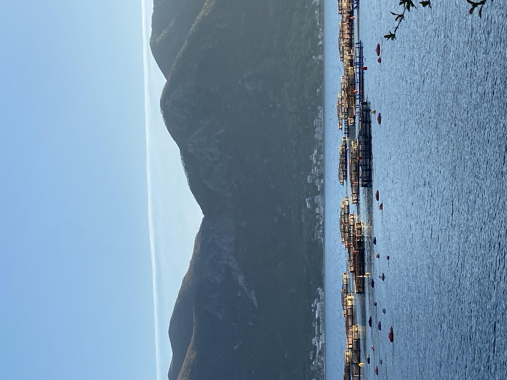 The image depicts the coastal area of Donji Orahovac, Montenegro. The scene shows dark, rugged mountains along the left side of the frame, transitioning into a narrow coastline with buildings. The right side of the image is dominated by the blue water of the bay. There are several structures that look like fish farms in the water, with red and yellow floats visible. A few branches are hanging over the water, suggesting the vantage point is from land. The sky is clear blue near the mountains.