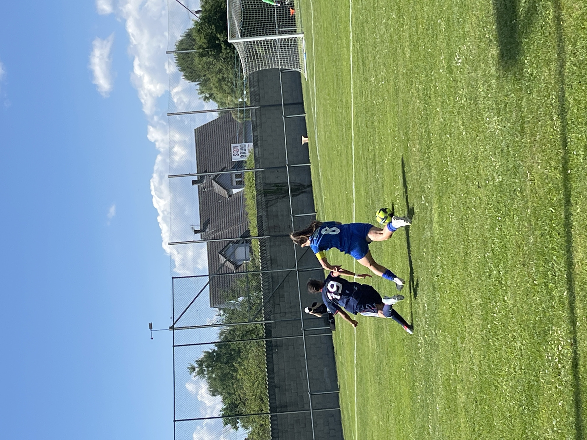 This full-frame photograph depicts an outdoor soccer match in progress under a clear blue sky with scattered white cumulus clouds, set in Porte du Ried, France.
In the foreground and midground, two female soccer players are actively engaged on a green grass field. The player on the left wears a dark blue jersey with white stripes on the sides and the number "19" in white on the back, dark shorts, and white cleats with red accents. Her dark hair is pulled back. She is in a forward motion, facing slightly away from the camera, moving towards the right side of the frame. The player on the right wears a blue jersey with the number "8" in white on the back and partial vertical text "TRICH" on the right side, blue shorts, and white cleats. Her brown hair is tied back. She is also in motion, positioned to engage with the ball. A yellow and black soccer ball rests on the grass between these two players, near the right player's left foot.
A tall chain-link fence with dark posts spans horizontally across the midground, backed by a light gray concrete block wall. Behind this wall and fence, several residential buildings with dark tiled roofs are visible amidst green trees and shrubs. A prominent sign, a white rectangular banner with "SODY" above "BRUNO" in dark letters, hangs on the fence structure in front of one of the houses. Above the fence line, near the center, a small, metallic, arrow-shaped wind vane is visible against the sky, pointing to the left.
Additional individuals include a person in the midground, partially obscured behind the concrete wall, wearing a white cap and dark clothing, holding what appears to be a camera. On the far right, a white-framed soccer goal with a net is visible, and a person in a green top is partially seen behind it. The scene is brightly illuminated by natural sunlight, casting discernible shadows on the field.