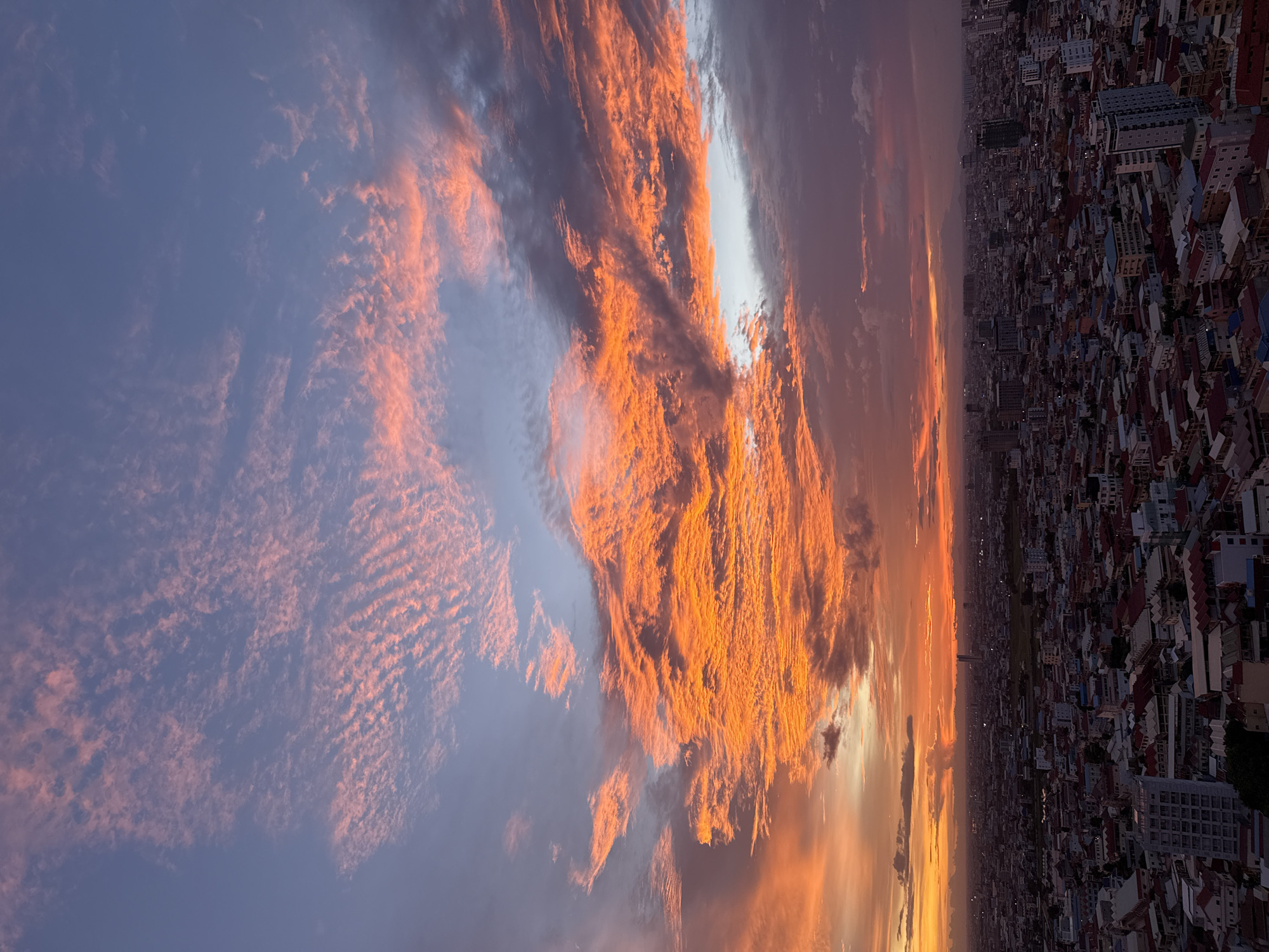 This is a vertical, wide-angle, aerial photograph of the city of Khan Mean Chey, Cambodia, at sunset. The sky dominates the upper two-thirds of the frame and is filled with a dramatic display of clouds. The clouds are predominantly orange and pink, illuminated by the setting sun, with streaks of lighter blue and gray visible. The cityscape occupies the lower third of the image. It shows a dense urban environment with many buildings of varying heights and architectural styles, predominantly residential with red-tiled roofs. Some taller, modern buildings, likely offices or apartments, are interspersed among the lower structures. The overall lighting is dim, characteristic of twilight, with the bright clouds in the sky creating a stark contrast with the subdued tones of the city below. There are no visible people or vehicles in the frame. The photograph captures a moment of natural beauty over an urban expanse.