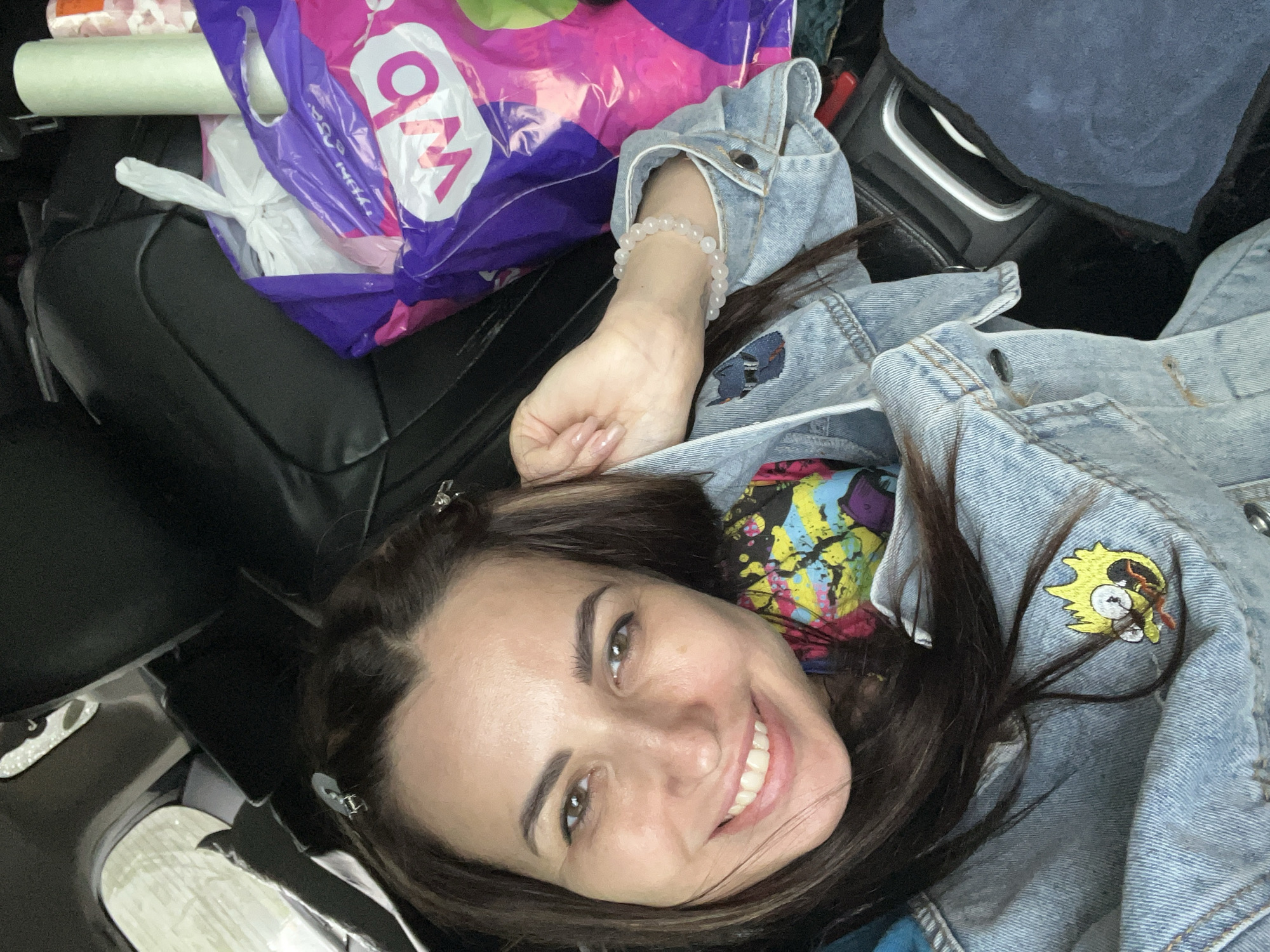 A selfie of a person with fair skin and dark brown hair, who is smiling at the camera. They are wearing a light blue denim jacket, under which a colorful patterned garment is visible. A white bead bracelet adorns their left wrist. The person appears to be inside a vehicle, as a black leather seat is visible to the left, and part of the car's interior is visible in the background. There are shopping bags and a roll of packaging material on the seat next to the person. Location: Unknown, Unknown.