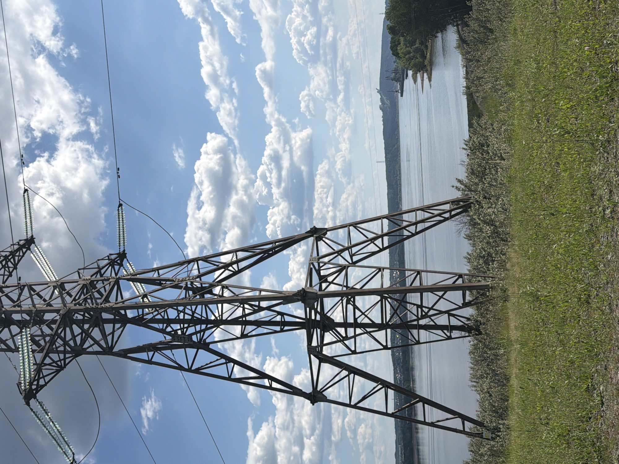 This image depicts a large metal power line tower situated on a grassy hill overlooking a wide river under a partly cloudy sky. The tower is in the foreground, dominating the left side of the frame. It is constructed from dark, weathered metal beams forming a lattice structure. Several power lines extend from the top of the tower across the frame.
In the midground, the calm, gray water of the river stretches across the width of the image. On the far bank of the river, a tree-lined shore is visible, with a settlement and industrial structures, including cranes, dotting the landscape. The horizon is a gentle curve of hills.
The sky above is a vibrant blue, dotted with numerous fluffy white clouds, suggesting fair weather. The grassy hill in the foreground is covered in green grass and some lighter-colored scrub vegetation. There are no people or animals visible in the scene. The overall impression is one of an industrial landscape integrated into a natural setting. The image appears to be taken in or near the city of Noviy, Russia, based on the architectural style of the industrial elements and the geographical features.