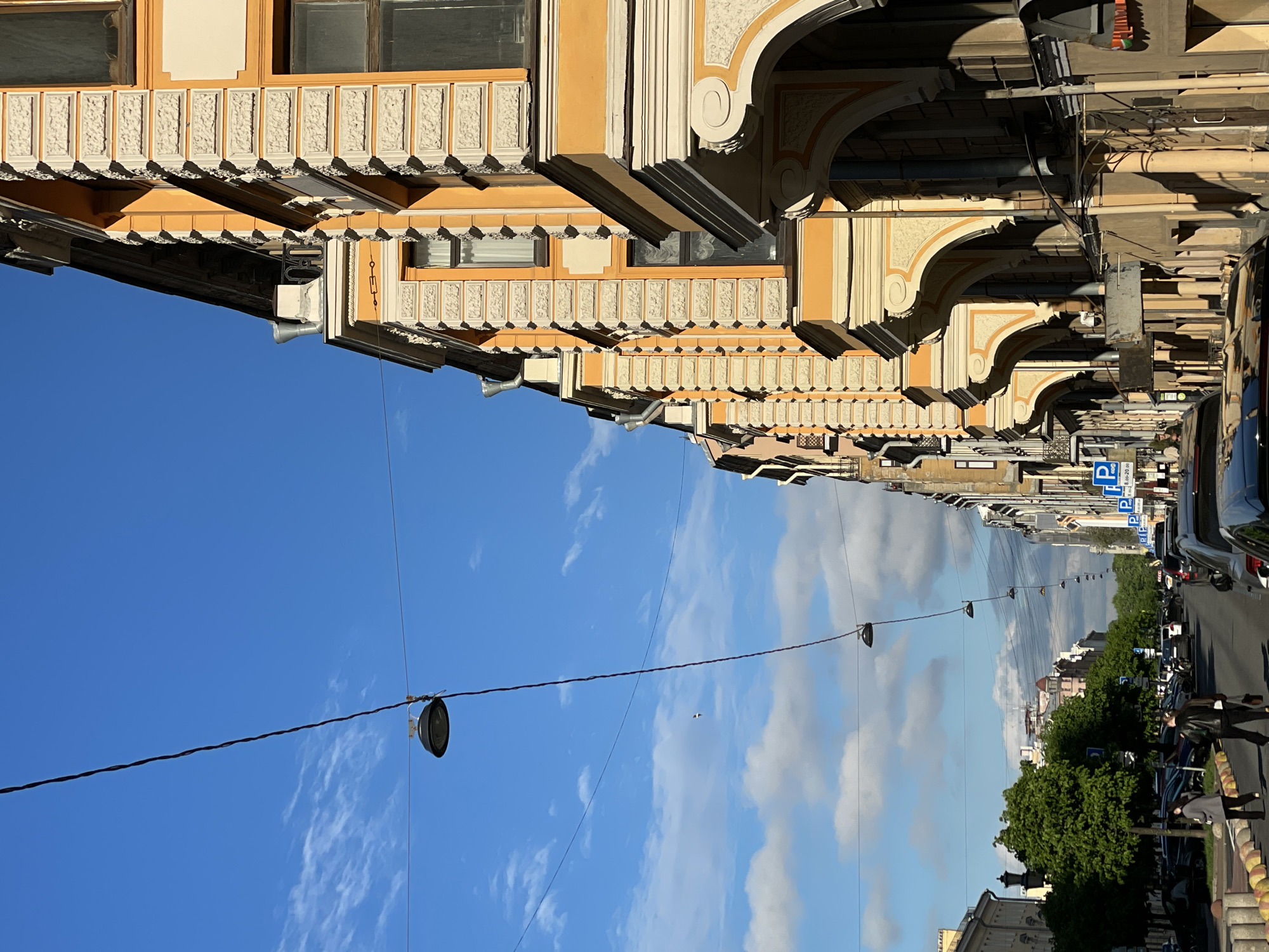 The image depicts a street scene in Saint Petersburg, Russia, captured from an unusual, near-vertical angle. Ornate, yellowish-tan buildings with detailed architectural moldings line the upper portion of the image, suggesting a historic district. The lower half of the image shows the sky transitioning into a street view. A clear blue sky dominates the left side, gradually shifting into clouds reflected in the road surface. Suspended overhead wires with streetlights run the length of the street. Cars are parked along the right side of the road. Trees and additional buildings are present along the street, leading into the distance. Pedestrians are walking on the sidewalk to the right. Parking signs are visible along the street.