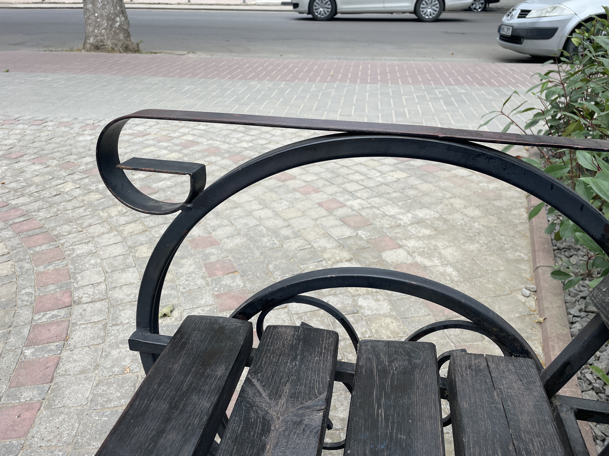 An outdoor perspective shot focuses on a section of a dark wooden slatted bench, its metal frame featuring curved scrollwork and a horizontal armrest. The bench is positioned on a patterned sidewalk composed of grey and reddish-pink rectangular tiles arranged in a semicircular fashion. Beyond the sidewalk, a paved street is visible, with two cars present: one a light-colored sedan in the foreground, and another lighter-colored vehicle further back. A tree trunk is partially visible on the left side of the frame, with its bark textured. Green foliage from a bush is seen on the right edge of the image, alongside a section of gravel. The scene appears to be a public urban area, likely in the city of Cahul, Moldova.