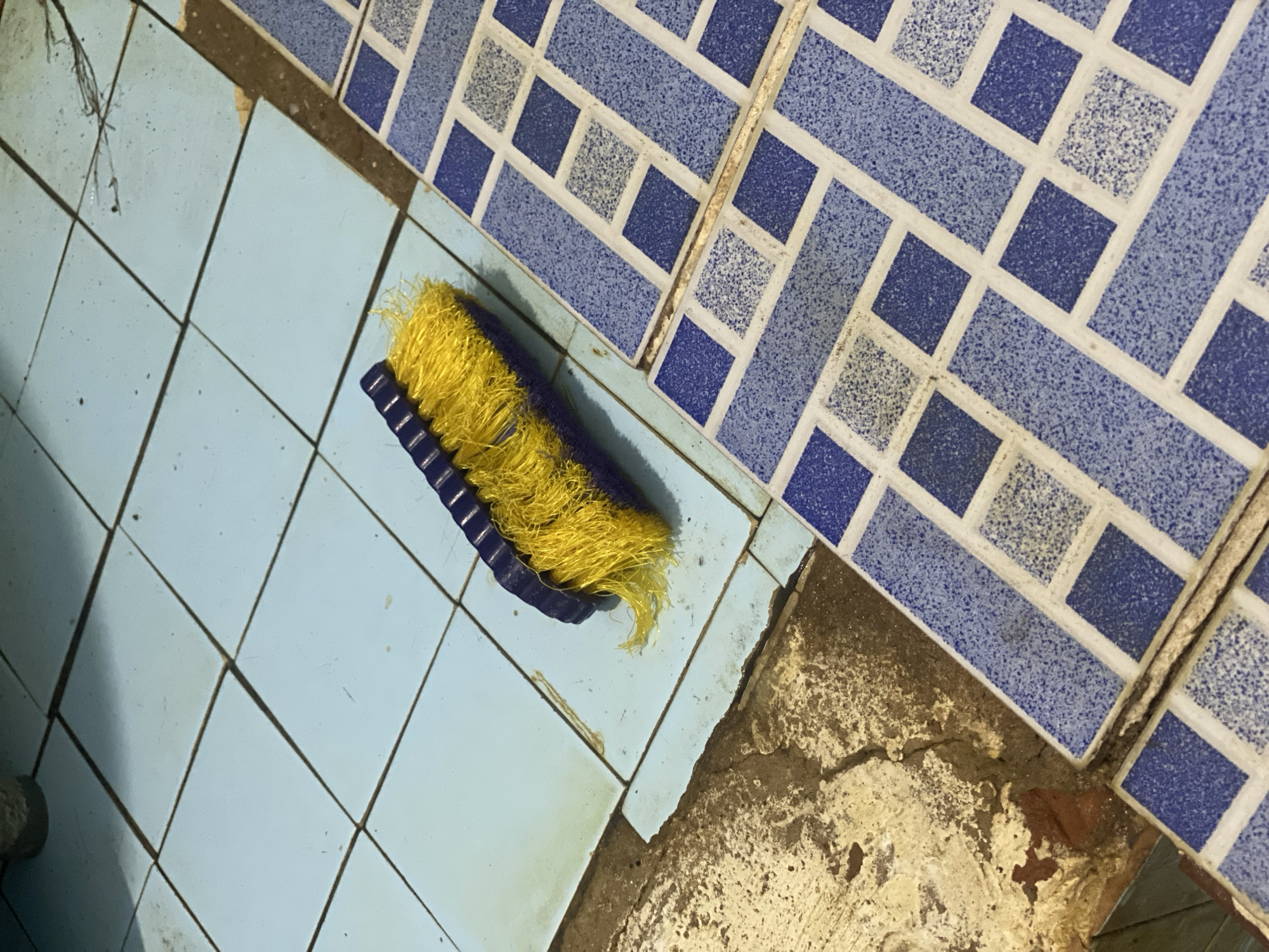A yellow and purple scrubbing brush rests on a light blue tiled floor. The floor is partially covered by a section of blue and white patterned tiles, and to the left, the tiled surface transitions into rough, uneven concrete. The brush appears worn, with some of its yellow bristles splayed. The scene depicts a mundane setting, possibly a bathroom or utility area, in Banjarbaru, Indonesia, given the context provided.