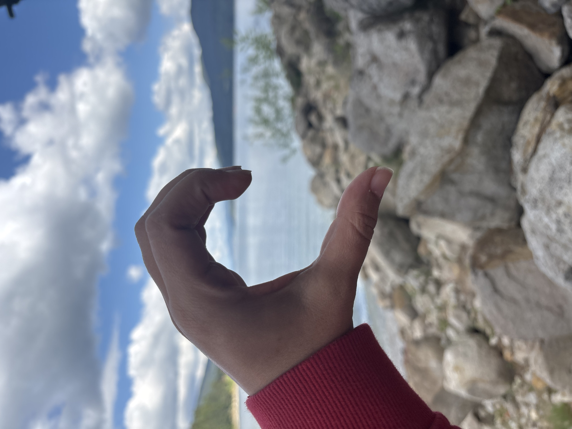 A person's hand, wearing a red sweater, forms the left half of a heart shape against a blurred background of a lake, mountains, and clouds. The hand is positioned in the foreground, with its fingers curved and thumb extended. The background shows a body of water, with distant mountains and a sky filled with white, fluffy clouds. The ground in front of the hand is covered with rocks and pebbles. The image is taken from a low angle, looking upwards towards the sky.