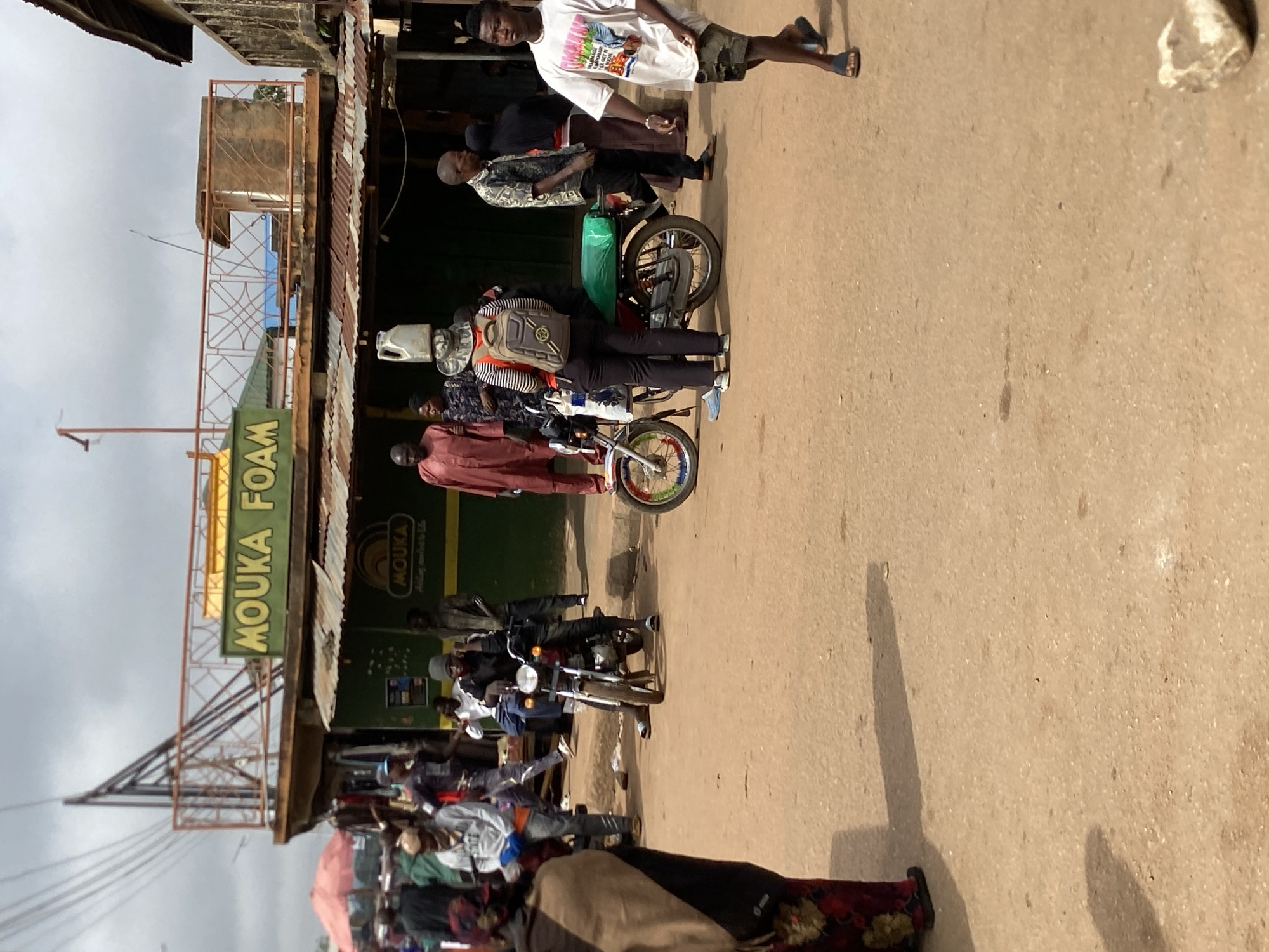 The image depicts a street scene in Bukuru, Nigeria, with a building marked "MOUKA FOAM" on the left side of the frame. Several people are present; some are walking, while others are riding motorcycles. The street is unpaved and has a brown, earthy tone. The sky is overcast. In the upper part of the picture, a man wears a white shirt and stands next to another man on a motorcycle with a green bag attached. Further down the street, a man in a maroon jacket is seen standing near another motorcycle. The overall environment is that of a busy urban street in Bukuru.