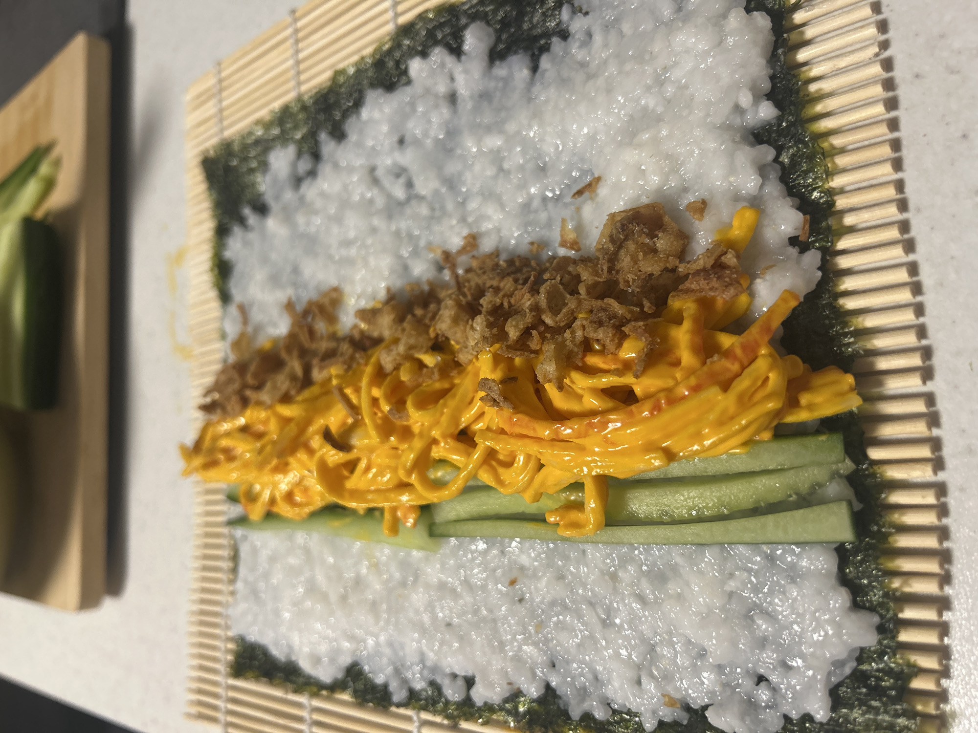 The image shows ingredients for sushi being assembled on a bamboo rolling mat. Nori seaweed is the base, covered with a layer of white rice. On top of the rice are cucumber sticks, a yellow sauce, and fried onion pieces. A cutting board with cucumber pieces is visible on the left side of the image. The picture was taken indoors. It is impossible to determine the location in the city of Veliko Tarnovo, Bulgaria, or any specific location without additional information.