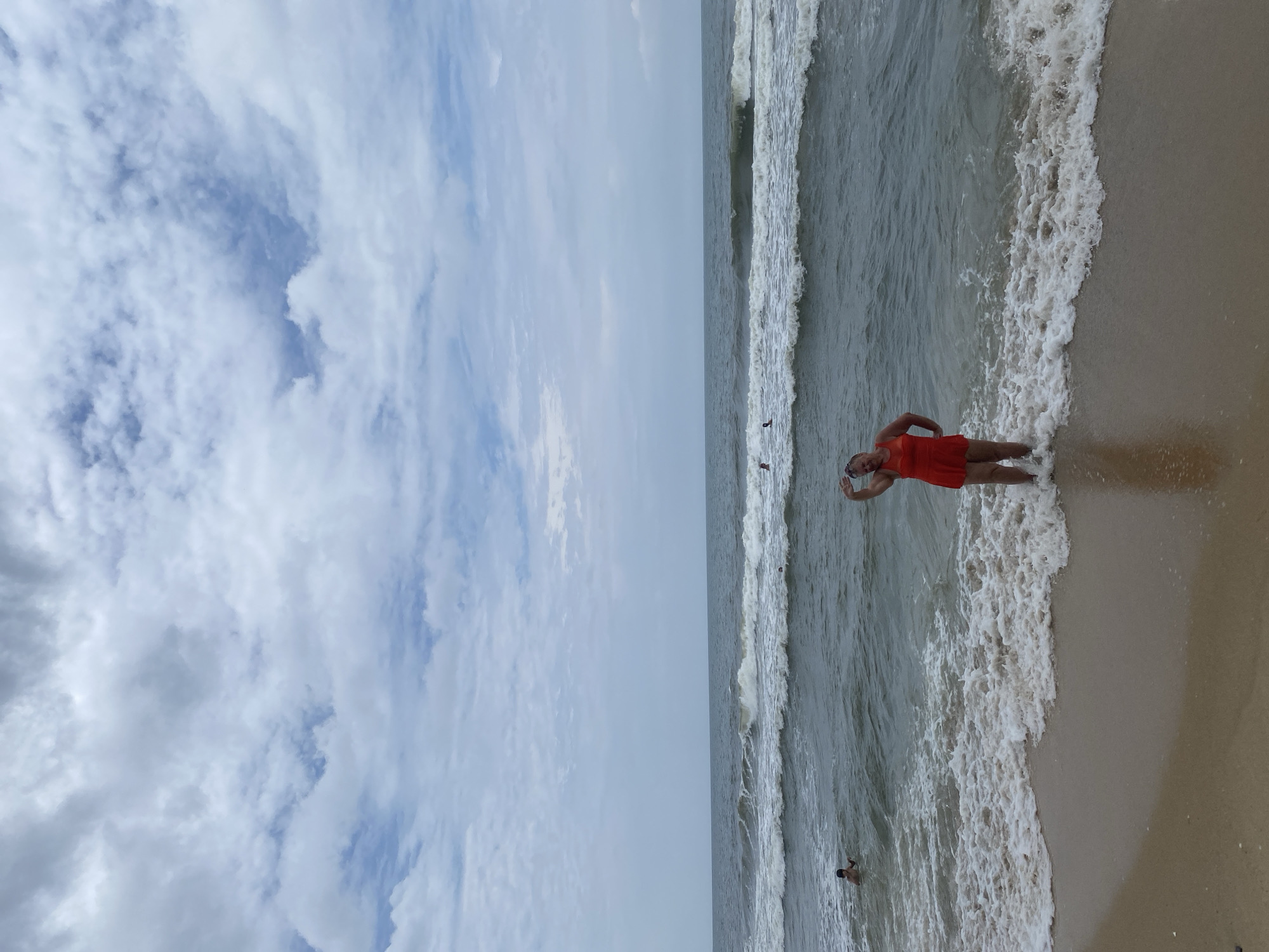 A woman wearing a bright orange swimsuit stands in the shallow water at the edge of the ocean, with waves gently washing over her legs. She is facing the camera, with her right hand raised to her forehead as if shielding her eyes from the sun, though the sky is overcast with scattered clouds. The beach is composed of light brown sand, and the ocean water is a muted green-grey color with white foam from the waves. In the background, several other people are visible in the water, some appearing to be swimming or wading. The horizon line is visible where the ocean meets the sky. The overall scene depicts a casual day at the beach in Mũi Né, Vietnam.