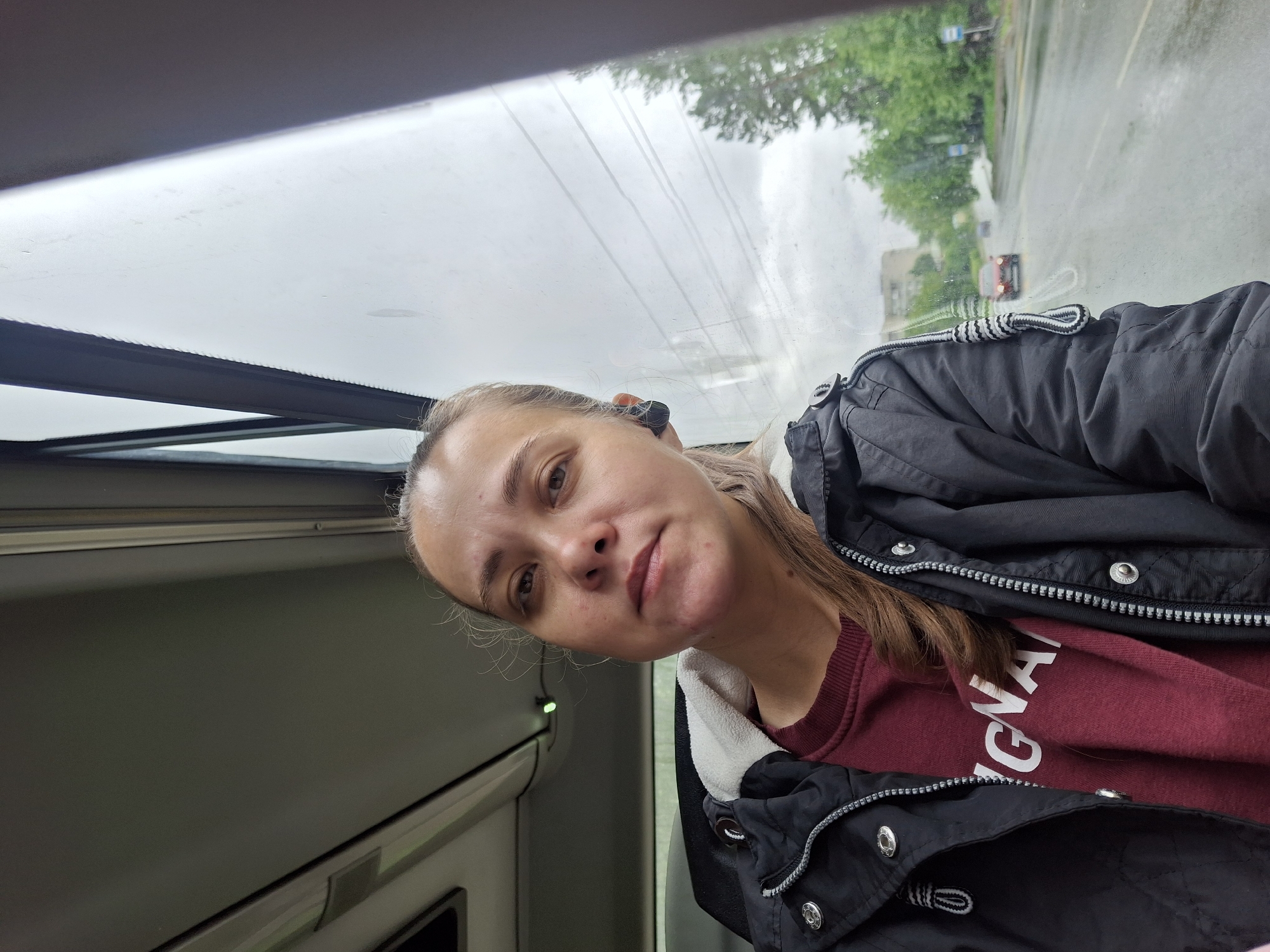 A woman is depicted in a medium close-up, situated inside a public transit vehicle, likely a bus, in Chaikovsky, Russia. She has light brown hair, some strands loose around her face, and wears a black earbud in her right ear. Her attire consists of a dark-colored jacket with a visible white collar lining over a maroon sweatshirt with partially visible white lettering ("GNA"). Her gaze is directed straight at the camera with a neutral expression. Through the vehicle's window to her right, a wet two-lane road with oncoming traffic, including a red car, is visible. The roadside is flanked by green trees and distant buildings under an overcast sky, suggesting recent rainfall. Reflections and horizontal lines, possibly from the window's heating elements or internal structure, are visible across the glass. The interior includes a grey overhead panel and part of a dark seat or partition.