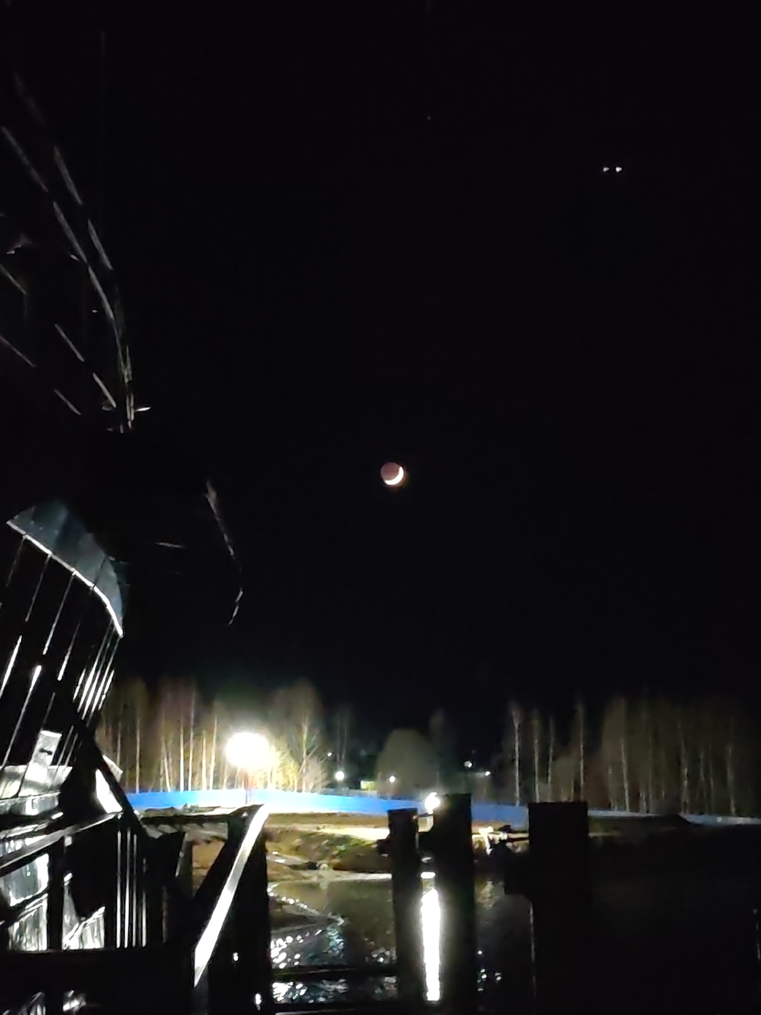 The image shows a view at night in Bol'shie Mkhi, Russia. A partial lunar eclipse is visible in the dark sky. Below the moon, there are trees illuminated by a light source. In the foreground, there appears to be a dark structure, possibly part of a building or bridge, reflected in the water. Two small bright objects are visible near the top-right corner of the image.