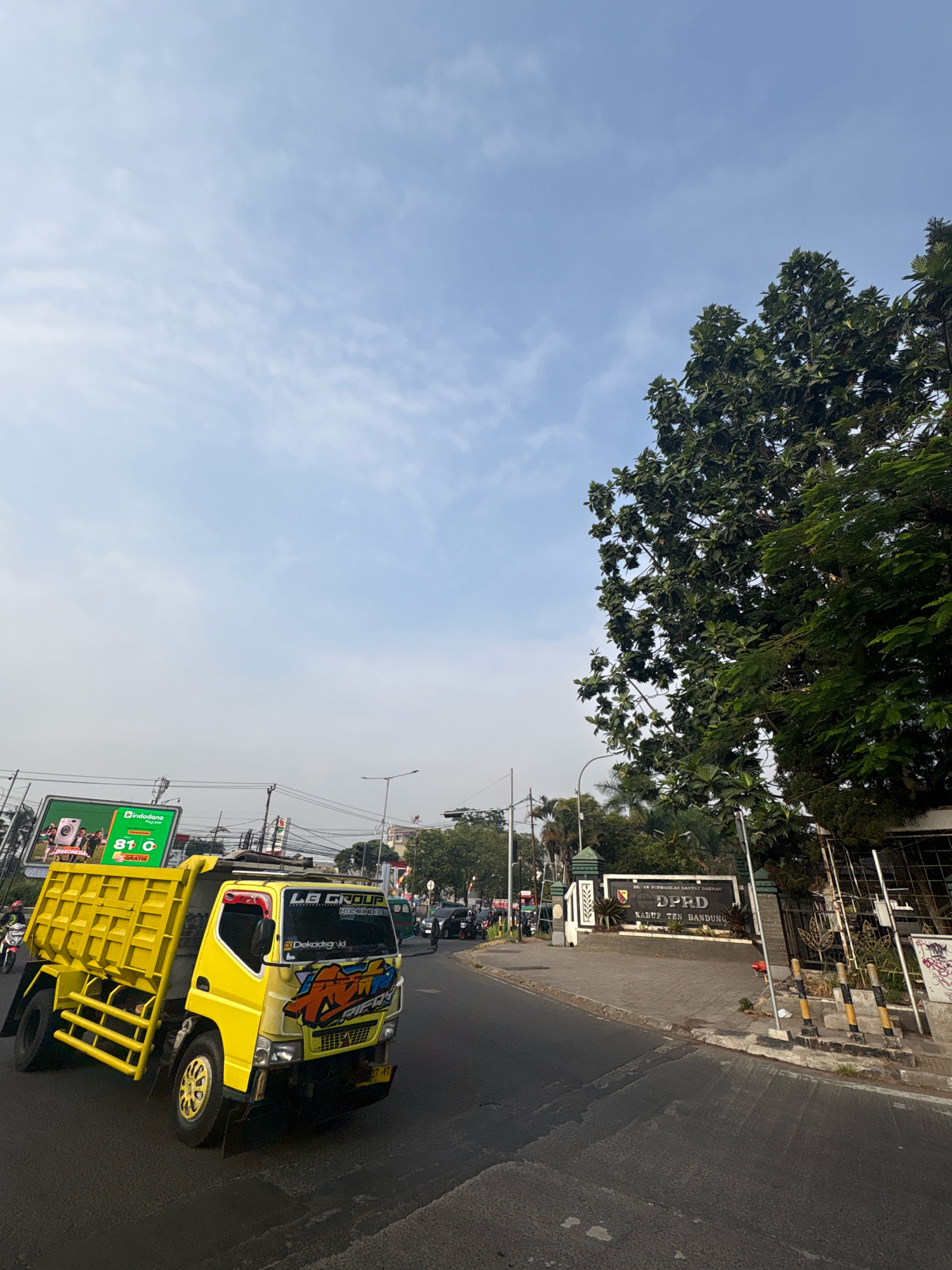 In Pamekaran, Indonesia, a bright yellow dump truck is captured in motion on a paved road. The truck features prominent branding with the text "LB GROUP" and stylized graphics on its front. The road is lined with a sidewalk, a decorative metal fence, and a sign that reads "DPRD KABUP TEN BANDUNG" (Regional People's Representative Council of Bandung Regency). A large tree with dense green foliage stands to the right of the sign, casting shadows. Overhead, a pale blue sky with wispy clouds dominates the upper portion of the image. In the background, other vehicles and traffic lights suggest a busy urban environment.