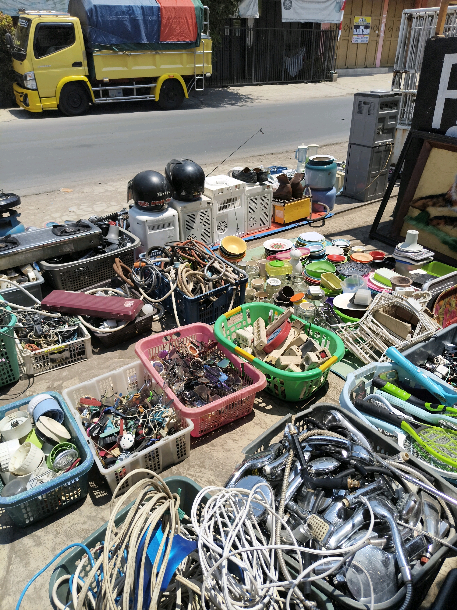 In Majasetra, Indonesia, on a sunny day, a street vendor has displayed a variety of used items for sale on the side of a road. The items are arranged on the ground and in plastic crates. In the foreground, a large gray bin is filled with numerous chrome-plated shower heads and hoses. To its right, a blue bin contains several electric mosquito swatters. Behind these, a pink basket overflows with eyeglasses, and a white crate holds a jumble of small electronic items and watches. Further back, a green basket contains what appear to be electrical components and adapters. Another collection of items includes various kitchenware, such as plates, bowls, and a blender, as well as a portable gas stove. Two black motorcycle helmets rest on top of white electronic devices, possibly air coolers. A yellow truck with a covered bed is parked behind the vendor's stall, and a metal fence encloses a building in the background. The scene suggests a typical street market environment.
