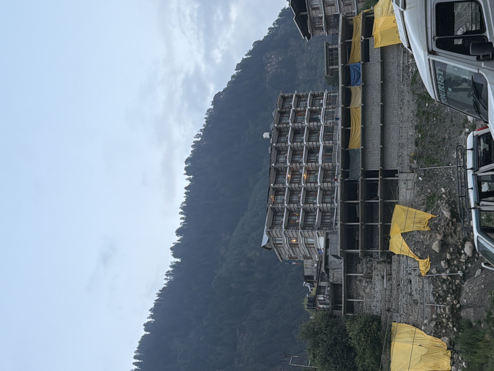 The image shows a multi-story hotel building constructed with stone and wood in Manali, India. The hotel is situated on a mountainside, with a dense forest covering the slope behind it. In the foreground, there are several vehicles, including a white van with "TOURIST" written on its windshield. Yellow tarps are visible in front of some of the lower levels of the hotel, possibly for construction or protection. The sky is overcast with clouds. The scene appears to be a developing area or a construction site, with unfinished structures visible below the main hotel building.
