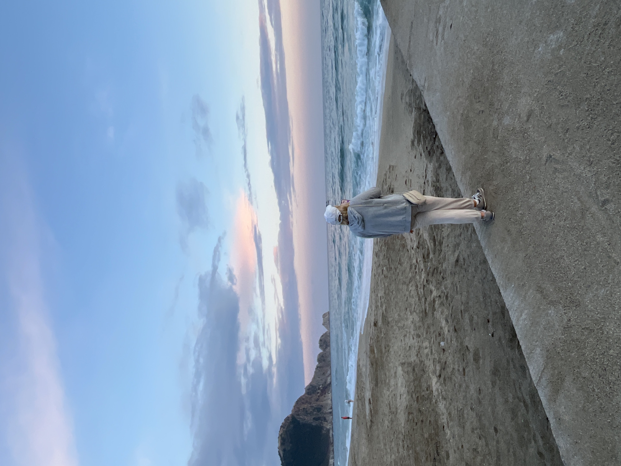 A person in light-colored clothing stands on a concrete structure overlooking a beach at Alanya, Turkey. The person is facing away from the viewer, towards the sea. To the left is a mountainous landform that projects into the sea. The sky transitions from a blue hue with scattered clouds to a pinkish-orange near the horizon. Waves are lapping onto the sandy shore.