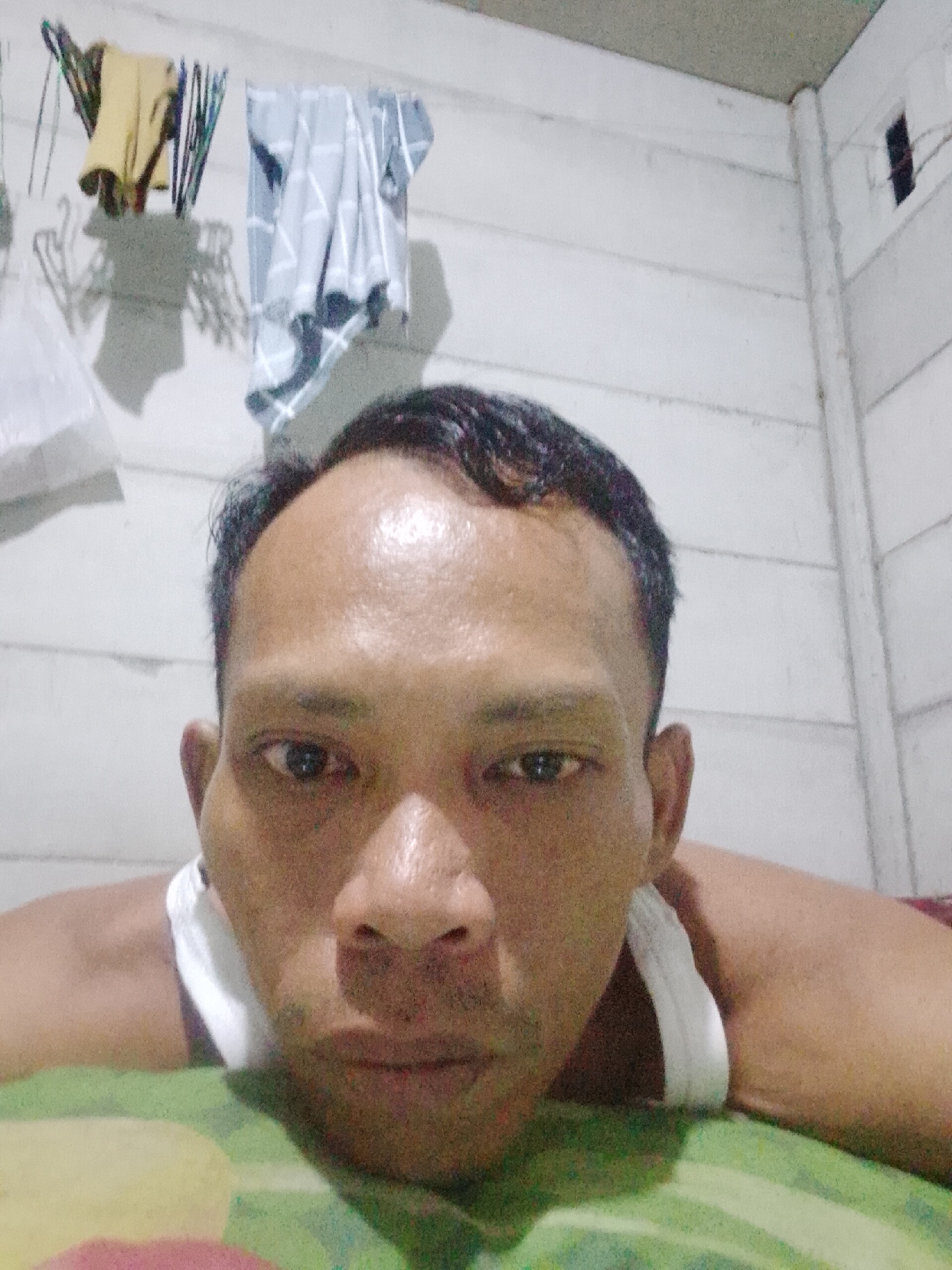 A man is positioned in the foreground, seemingly lying down, with his head closest to the camera. He has short, dark hair, and his face is the primary focus. He is wearing a white tank top.
The background shows a wall with horizontal panels, possibly made of wood. Clothes hang on hangers attached to the wall.
The scene appears to be indoors. It is impossible to confirm that the location is specifically Bukit Ketok, Indonesia, without further information or landmarks visible in the image.
