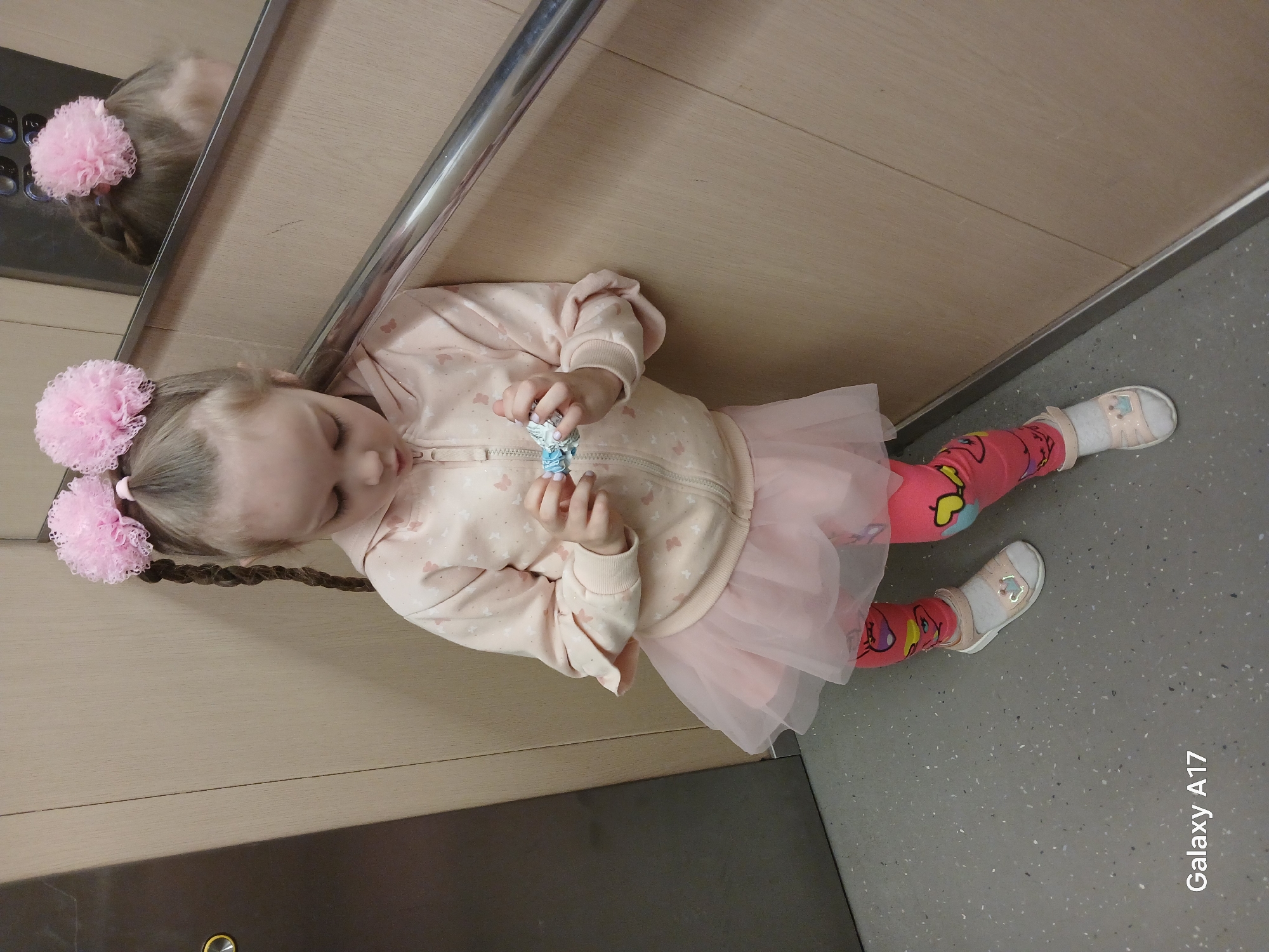 A young girl in a pink outfit with matching pink pompoms in her braided hair is lying on the floor in what appears to be an elevator in Yekaterinburg, Russia. The elevator has wood paneling and a silver handrail. The girl is holding a small blue and white object in her hands. She is wearing pink leggings with cartoon designs, white socks, and white sandals. Her reflection is visible in the elevator mirror. The floor of the elevator is gray speckled. The text "Galaxy A17" is visible in the bottom right corner of the image.