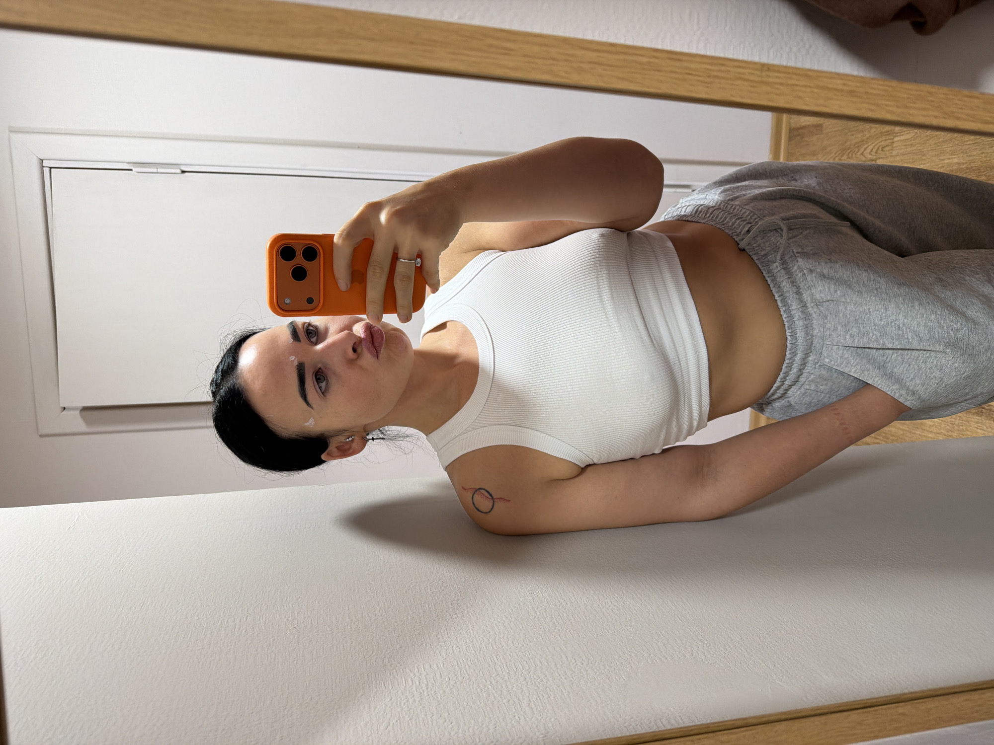 A young woman with dark hair, pulled back, is featured in a mirror selfie taken indoors. She holds an orange smartphone with a triple camera setup to capture her reflection. She is wearing a white ribbed crop top that exposes her midriff and a pair of grey sweatpants. A ring is visible on her left ring finger. On her upper left arm, there is a small tattoo depicting a black circle intersected by a red line. Her expression is neutral, with slightly pouting lips. There are small white marks on her forehead and cheek, possibly from a skincare product. The background, reflected in the mirror, shows a simple white wall or door, topped by a wooden frame or piece of furniture. The woman appears to be leaning or sitting on a light-colored, textured surface. No text is visible in the image. The photo is oriented sideways, rotated 90 degrees clockwise.
