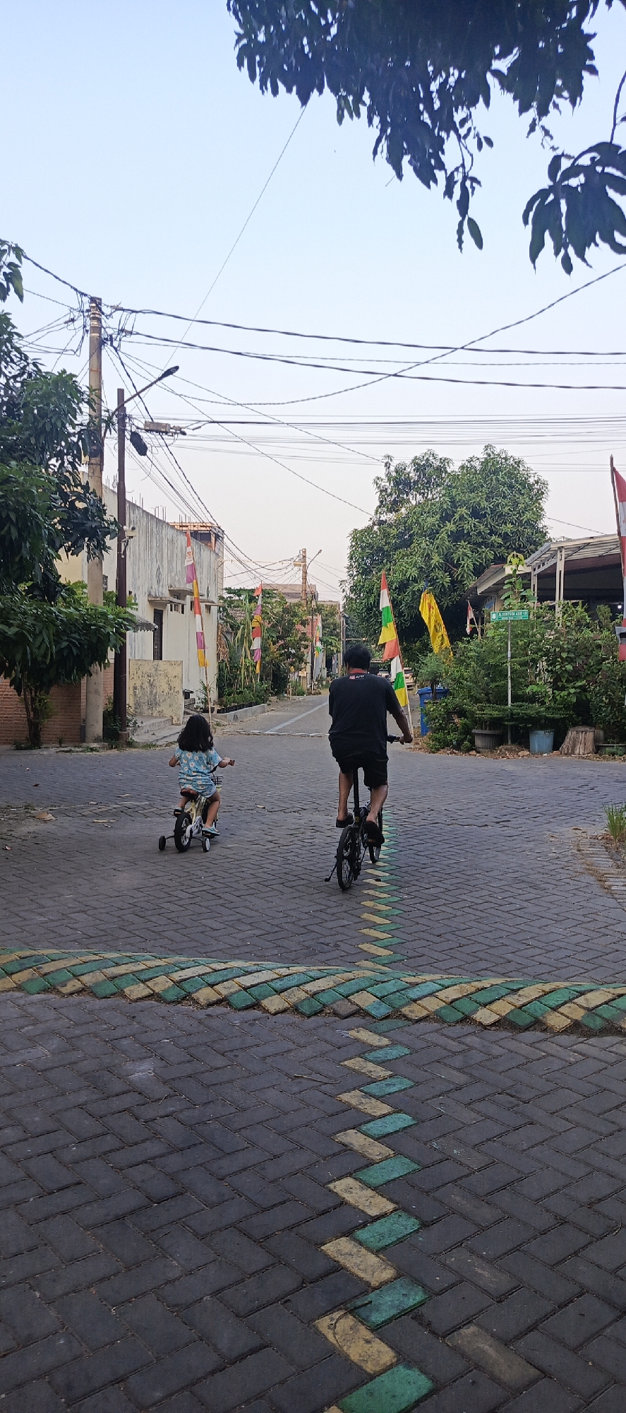 A full-shot, outdoor, eye-level, vertical, medium-angle photograph depicts a father and child biking down a paved street in Sukaresmi, Indonesia. The street is made of dark gray interlocking pavers, with a zigzagging pattern of yellow and green pavers in the foreground. The father, wearing a black t-shirt and shorts, rides a small black bicycle with a folding frame, his back to the camera. To his left, the child, with long dark hair, wears a light blue and white patterned t-shirt and rides a yellow tricycle with training wheels. The street is lined with buildings on both sides, including a light-colored house with a corrugated metal roof on the left and more distant structures with trees and vegetation. Indonesian flags in red and white and green and yellow are visible on poles along the street. Electrical wires crisscross the sky, which is a pale blue with a slight haze. Trees with dark green leaves frame the top of the image.