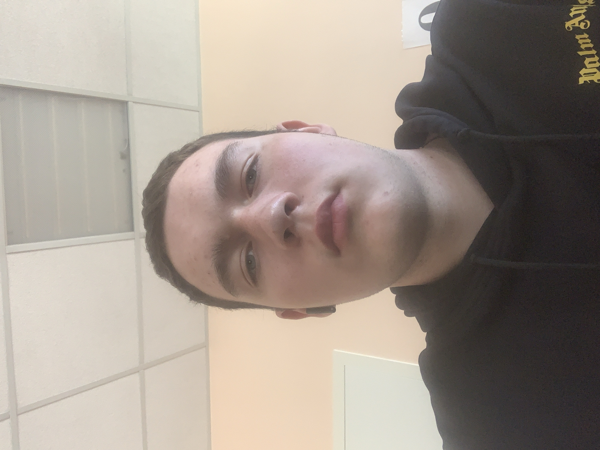 A close-up, selfie-style image shows a young man with short, dark hair and light skin. He is wearing a black hooded sweatshirt with a yellow logo on the shoulder. His face is angled slightly to the side, showing his nose, lips, eyes, and eyebrows. The background consists of a white, paneled wall and a light beige or peach-colored wall. A white door is partially visible in the background. The image's composition suggests it was taken indoors, possibly in an institutional setting like an office. There is no information to suggest a location in Severodvinsk, Russia.